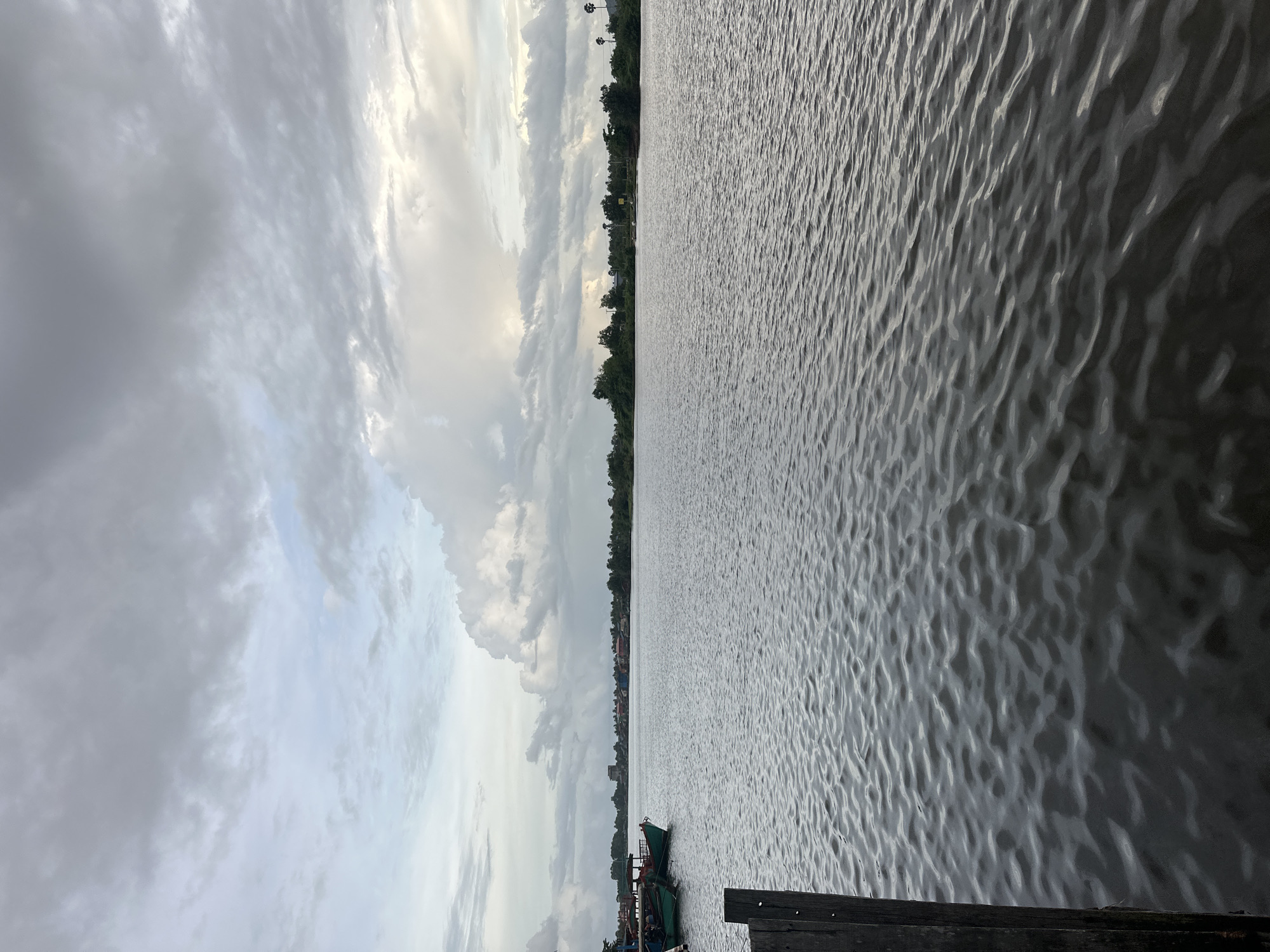 The image depicts a wide river with calm, choppy water under a cloudy sky. The sky is a mix of grey and white clouds, with hints of blue peeking through in some areas. On the left side of the river, a few green boats are docked near a wooden pier. The far bank of the river is lined with lush green trees and foliage, with some buildings visible amongst them, suggesting a populated area. The overall scene has a serene, somewhat overcast atmosphere. The location is the Kampot River in Kampot, Cambodia.