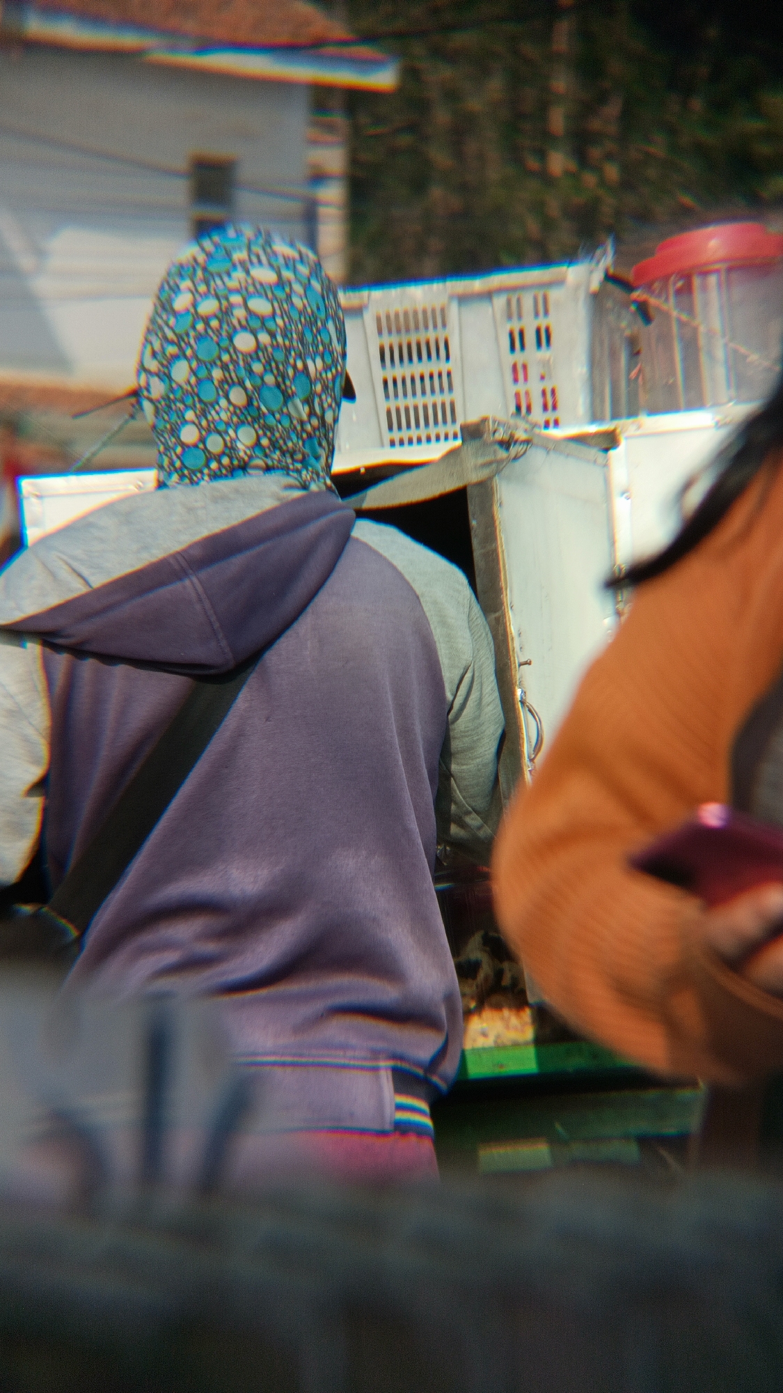 In Margajaya, Indonesia, a person wearing a blue and white patterned headscarf and a purple and grey hoodie is partially visible from behind. They are standing next to a white crate with ventilation slits, which is positioned on a metallic structure. To the right, another individual's arm, clad in an orange garment, is in the foreground, holding a smartphone. The background shows a blurred urban environment with buildings and greenery.