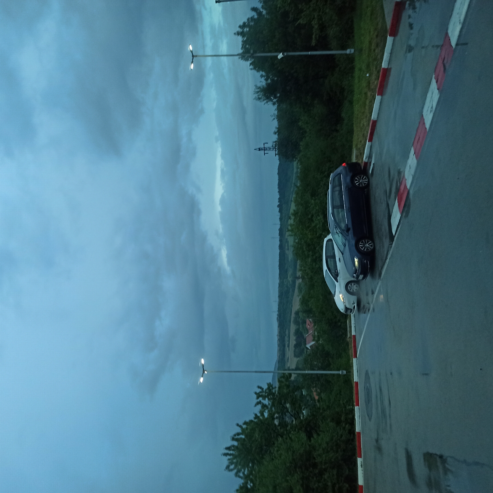 A view of a parking lot in Kralev Dol, Bulgaria, with two cars parked on a wet asphalt surface under a cloudy, overcast sky. The cars are a white hatchback and a dark blue station wagon. The parking lot is bordered by a red and white striped curb. Trees and vegetation line the background, with distant rolling hills visible under the dark clouds. Two tall lamp posts with multiple lights are present, one on the left and one on the right of the frame. A power line tower is also visible in the distance. The overall lighting suggests it is either early morning or late evening, or a heavily overcast day.