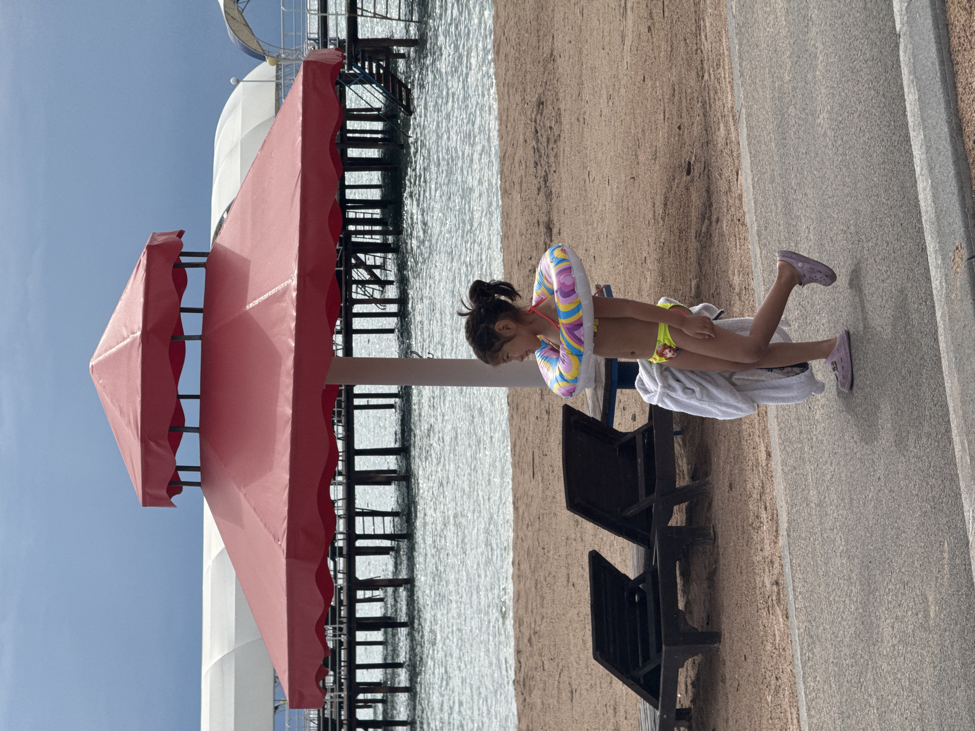 A young girl in a yellow bikini and a colorful inflatable ring around her waist stands on a sandy beach next to a covered seating area. She is wearing purple Crocs. The girl is looking down and appears to be preparing to sit on a dark brown lounge chair. A white towel is draped over the chair. To the left, a large red awning covers a portion of the scene. Beyond the beach, a body of water with a wooden pier is visible under a bright blue sky. The sun is shining, suggesting it is daytime. The scene is set in Sary-Oy, Kyrgyzstan, likely at a resort or beach area, given the lounge chairs and pier. There is no visible text in the image.