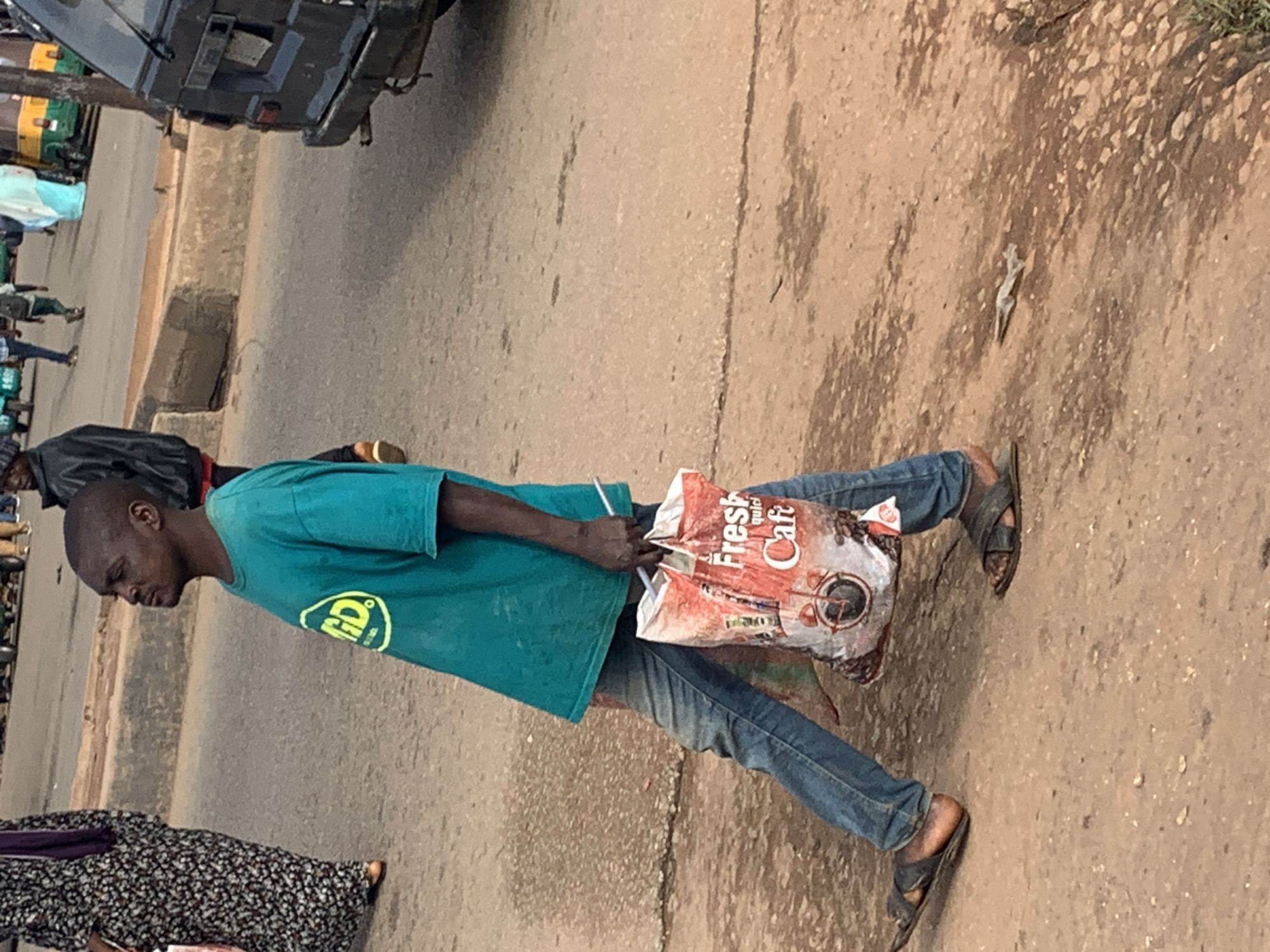 A man, appearing to be of African descent and wearing a teal t-shirt with a yellow circular logo, jeans, and dark sandals, walks from left to right across a dusty street. He carries a large, red and white plastic bag filled with what appears to be coffee, in his right hand. His gaze is directed to his left, away from the camera. To his left, a woman in a black and white patterned dress with a purple headscarf is also walking. In the background, a dark-colored van is parked on the right side of the street, and other vehicles and pedestrians are visible in the distance. The environment appears to be an urban setting, possibly a marketplace or busy street, with a curb on the left and uneven ground. The image was taken in Gura Top, Nigeria.