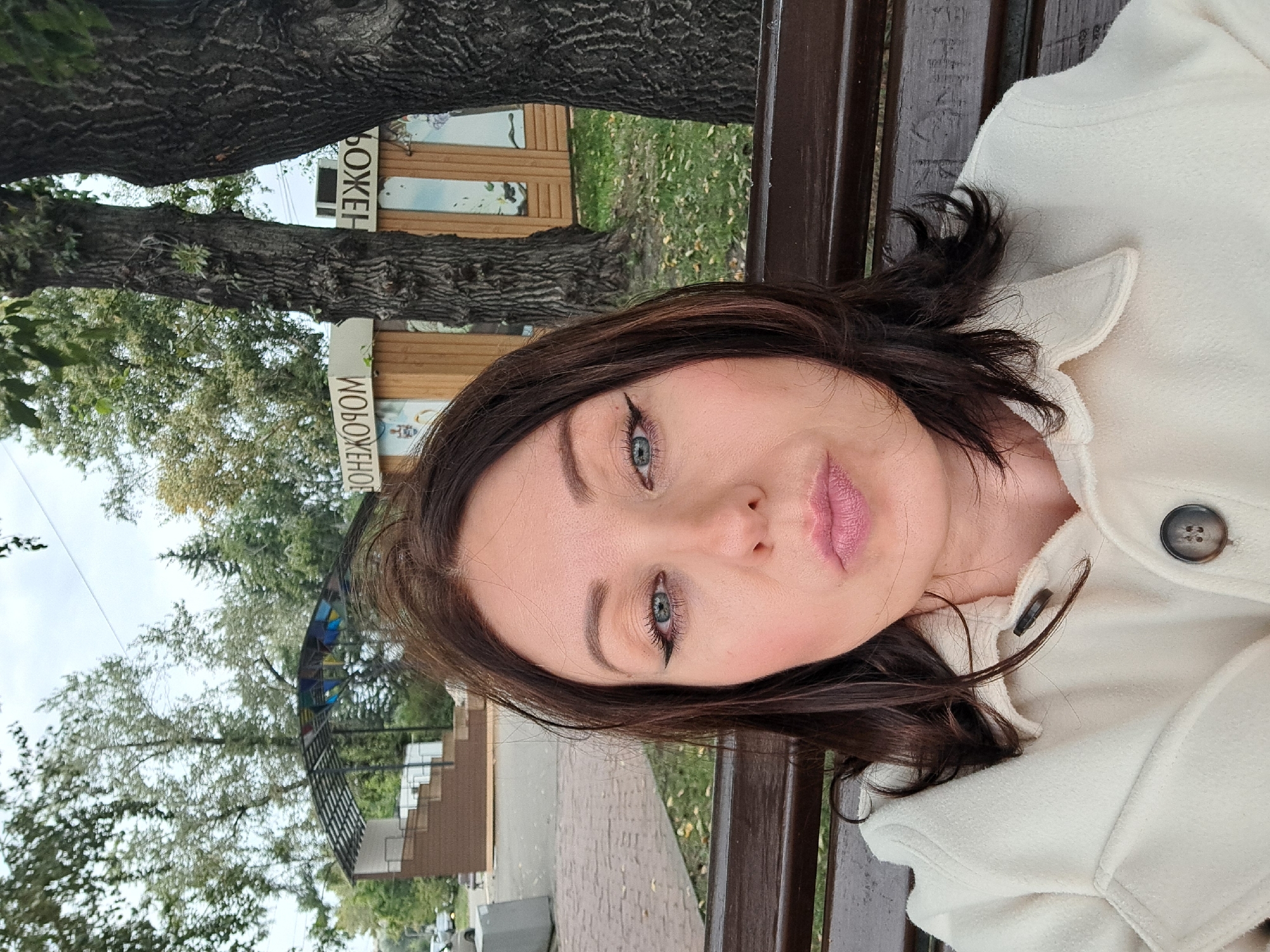 A close-up, waist-up selfie shows a woman with dark brown hair, light skin, and blue eyes, wearing a cream-colored jacket with a dark button. She is seated on a dark wooden bench, her lips pursed. Behind her, a park setting is visible with trees, grass, and a small open-air structure with tiered seating. In the background, a small wooden building with windows and signs that appear to be in Cyrillic script is visible. The signs read "МОРОЖЕНОМ," which translates to "ice cream." This suggests the location might be a park or public space in Irkutsk, Russia, possibly near an ice cream stand. The overall atmosphere is casual and outdoor.