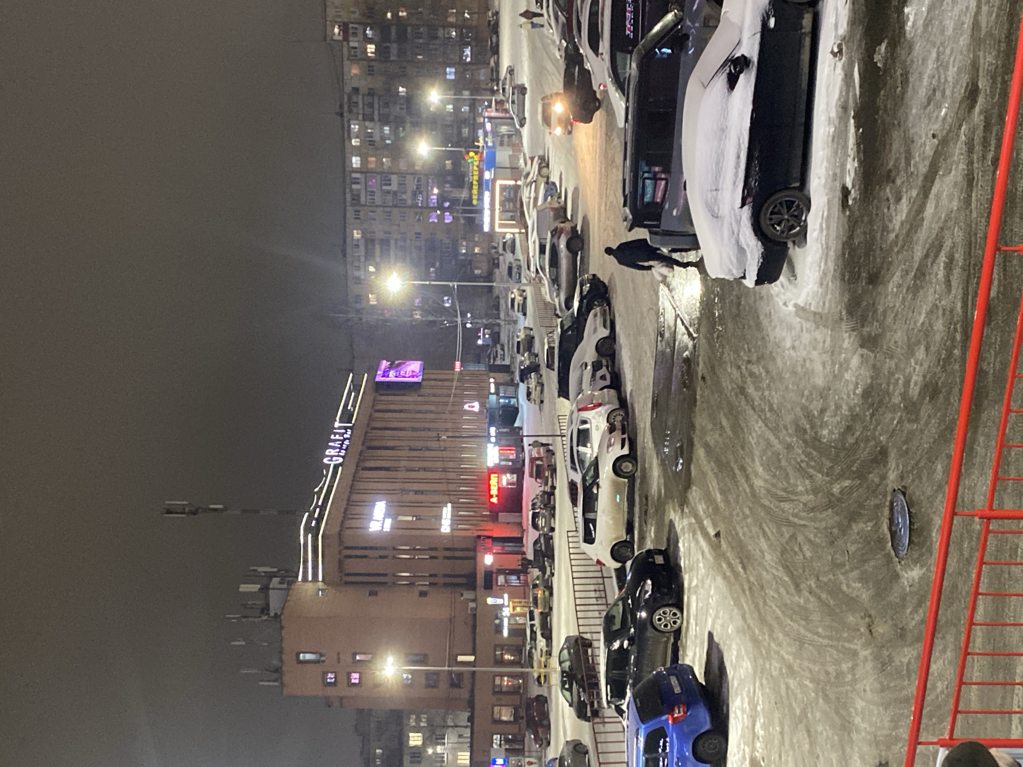 Here is a detailed description of the image:
**Overview:**
The image appears to be taken in the evening in an urban setting, likely in winter due to snow. It showcases a mix of buildings, parked cars, street lighting, and a generally cold environment.
**Location:**
Based on the visible signs, the location is in Izhevsk, Russia. There is a building with "GRAFIT" signage.
**People:**
There's at least one person visible near the cars, possibly walking or interacting with a vehicle.
**Objects and Subjects:**
* Multiple parked cars lined along a street or parking area. Some of the cars are covered in snow.
* Buildings, including residential blocks and commercial buildings with lit signs.
* Streetlights providing illumination.
* A red metal fence or barrier in the foreground, possibly near the photographer's location.
* Various signage on buildings, including "A-Irena."
* A manhole cover in the foreground.
**Actions and Interactions:**
* People walking among the parked cars.
* Cars are parked along the street.
**Environment:**
* The setting is an urban environment with a mix of residential and commercial buildings.
* It appears to be night.
* Snow is present, indicating a cold, possibly winter season.
* The lighting is a mix of streetlights and building illumination.
