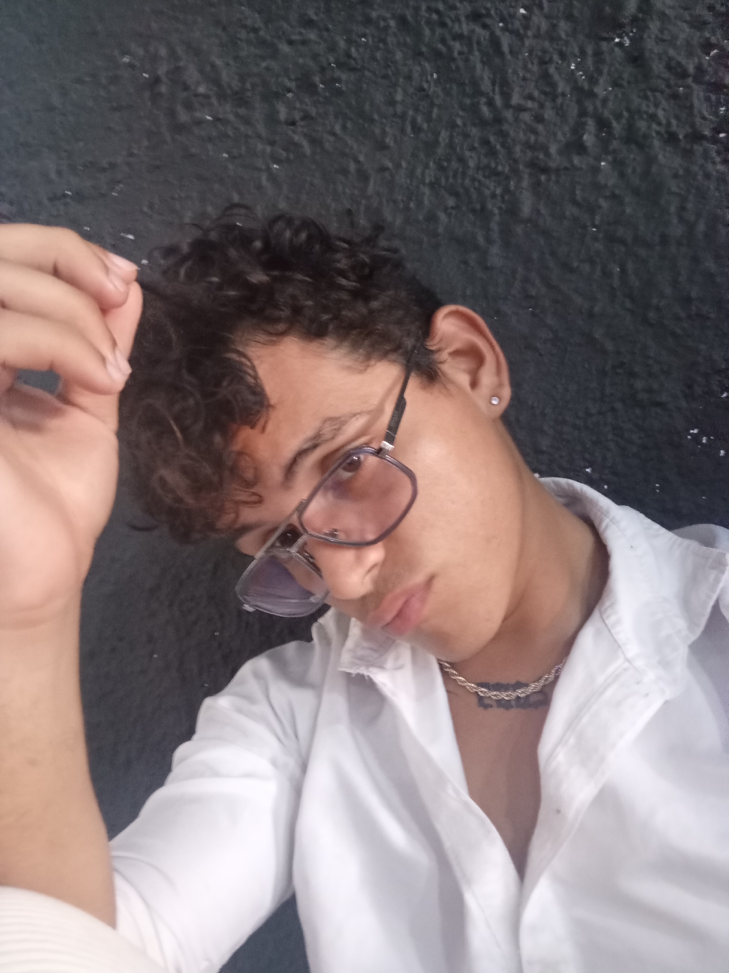 A person with curly dark hair and glasses is looking down and to the right. They are wearing a white collared shirt and a silver chain necklace. Their right hand is raised to their hair. The background is a dark, textured wall. The image is a selfie taken from a high angle. The location is not specified in the image.