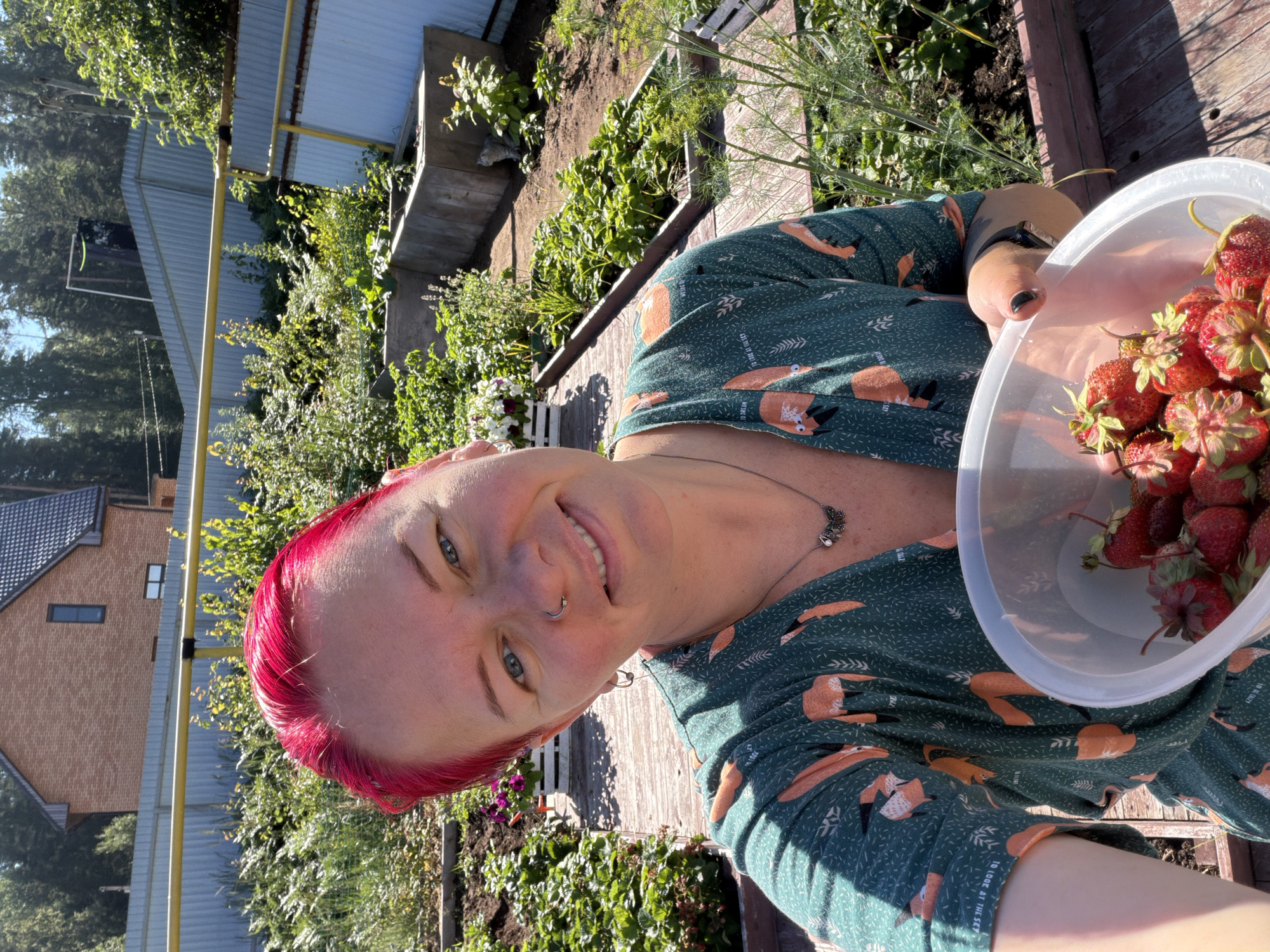 A woman with bright pink hair is taking a selfie in a garden. She is wearing a green dress with a fox print and is holding a clear plastic bowl filled with strawberries. The garden appears to be in a residential area, with a brick house and a metal fence visible in the background. The weather is sunny, and the plants are lush and green, suggesting it is summer. The woman is smiling at the camera, and her left arm is visible, holding the bowl. The image seems to be taken on a wooden deck or pathway within the garden. This scene is located in the city of Chaikovsky, Russia.