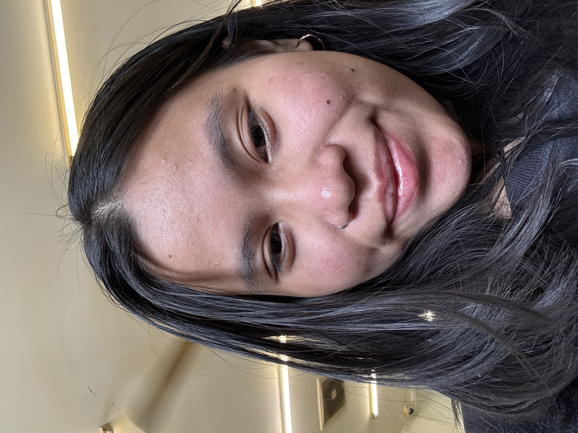 A close-up selfie shows a young East Asian female with dark hair, looking directly at the camera. She is wearing a black top, and a silver hoop earring is visible on her right ear. The background appears to be an indoor setting with a beige ceiling and fluorescent lighting fixtures. There are no distinguishing landmarks or objects in the background that would indicate a specific location within East Jakarta, Indonesia.