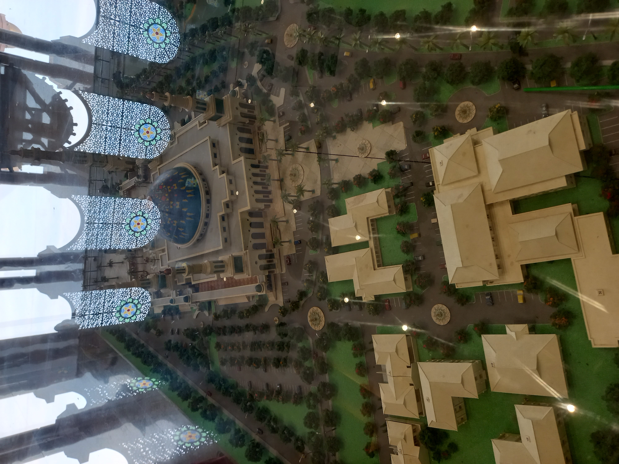 The image displays a detailed architectural model of a mosque and surrounding buildings, likely a university campus or religious complex, set within a miniature city environment. The primary structure is a large mosque with a prominent blue dome adorned with colorful patterns. The mosque features multiple minarets and a courtyard. Surrounding the mosque are several other buildings with light-colored roofs, designed in a modern architectural style. The model includes meticulously crafted landscaping with trees, grassy areas, parking lots with miniature cars, and pathways. Decorative elements, such as ornate screens with geometric patterns and circular fountains, are integrated into the design, suggesting a focus on aesthetic detail. The overall scene evokes a sense of order and planned urban development within a simulated environment. The model is likely situated indoors, as indicated by reflections on a transparent surface, possibly glass, covering the display. The scene is set in Samarinda, Indonesia.