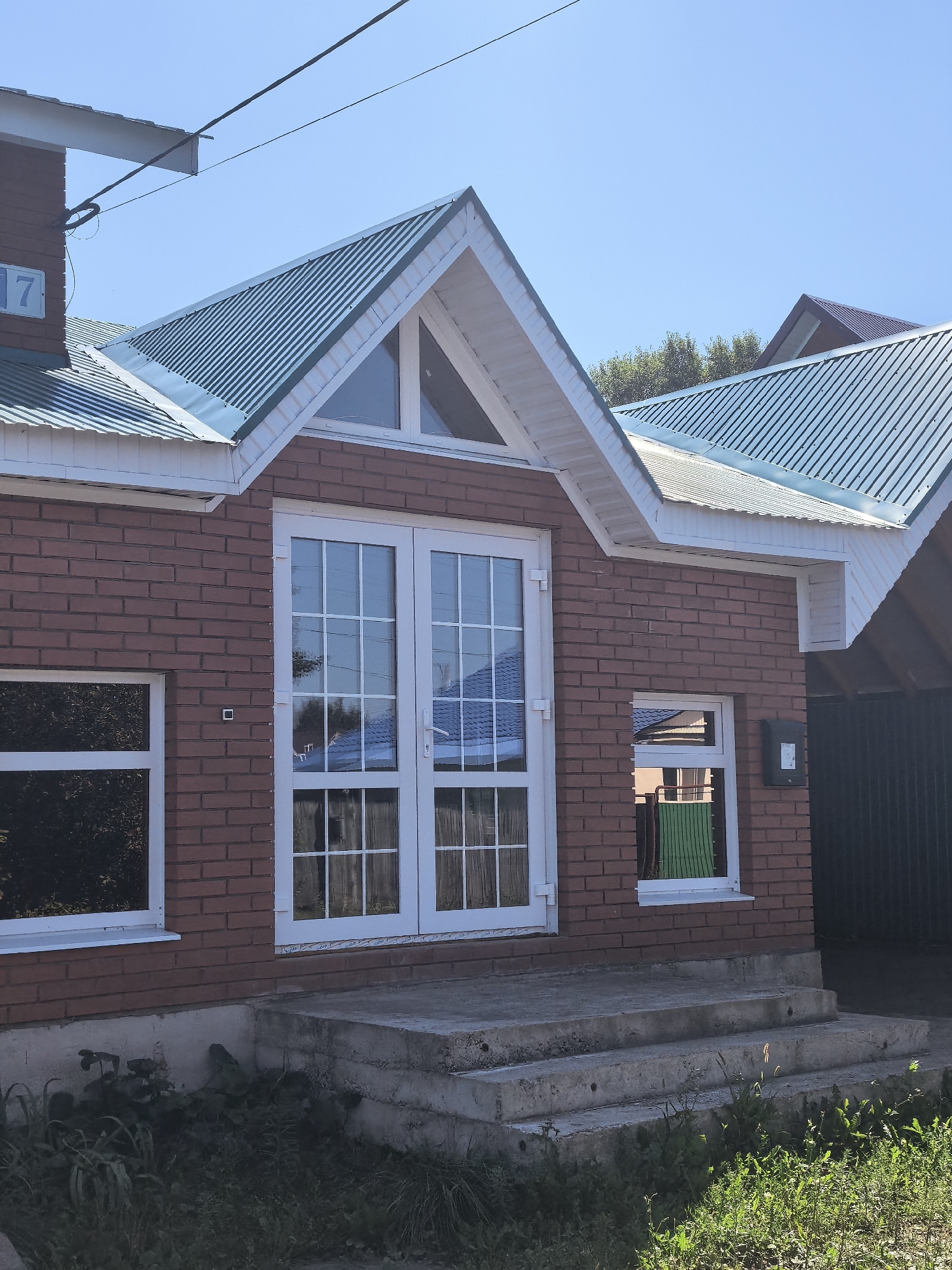 The image depicts the exterior of a two-story brick house in Sterlitamak, Russia, on a clear day. The house features a prominent green metal roof with white trim. On the left side of the facade, a white-framed window is visible, reflecting trees. Double doors with multiple panes, also framed in white, are centrally located, leading to a set of concrete stairs. To the right of the doors, another smaller white-framed window shows a green object inside. A black electrical box is mounted on the brick wall next to this window. The house is situated in a residential area, with other roofs and greenery visible in the background, and a dark fence on the far right. The foreground shows overgrown grass and vegetation around the base of the concrete stairs. A blue sign with the number "17" is attached to the upper left section of the house. Power lines are visible against the clear blue sky.
