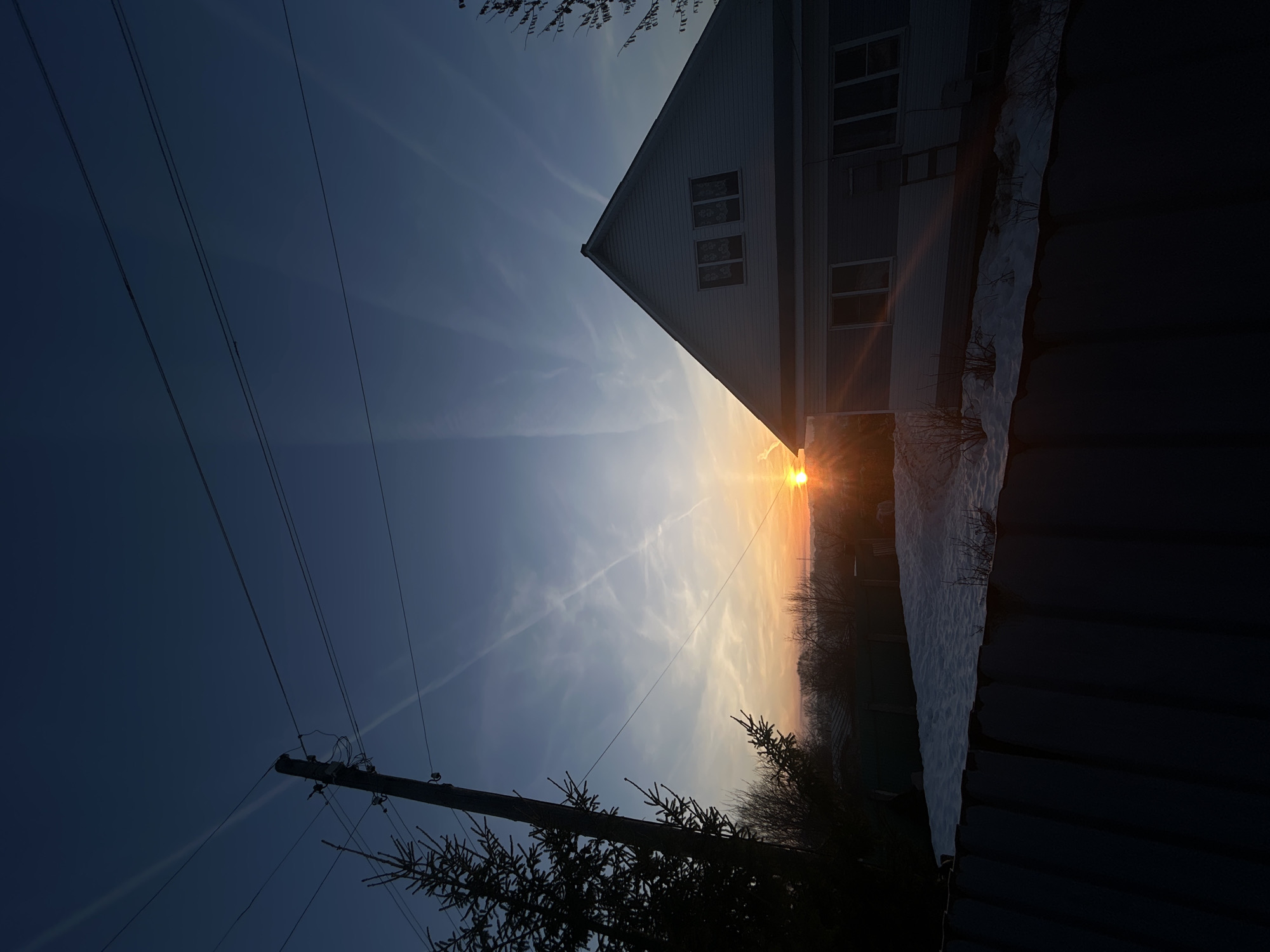 The image depicts a sunset view, likely in a rural or suburban area. A house with a gable roof is visible, partially obscured by a fence and some trees. The sun is setting directly behind the house, creating a bright glare. Power lines and a utility pole are present in the foreground. The sky is a mix of blue and orange hues. The presence of snow suggests a winter setting. The image is taken in the city of Фоки, Russia.