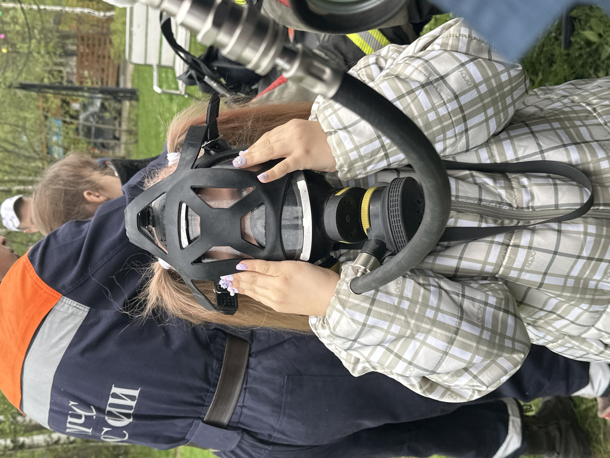 In Grebnevo, Russia, a person wearing a plaid jacket is shown putting on a black respirator mask with a black hose and yellow filter. The individual's hands are visible on the mask, adorned with light-colored floral nail art. To their left, a person wearing a dark blue uniform with orange and silver accents stands close. The uniform has white text visible on the left side. In the background, there is green grass, a tree, and a piece of silver equipment.