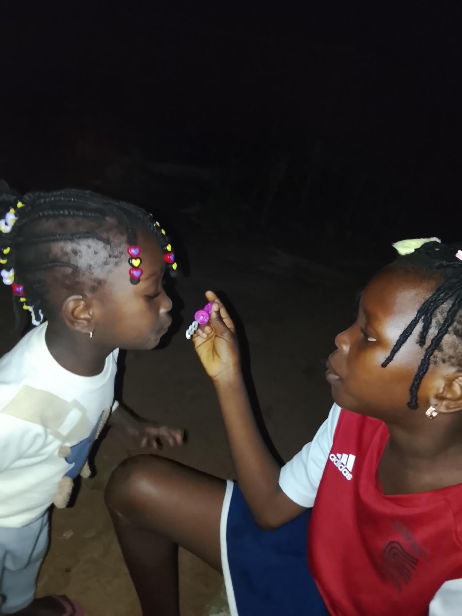 Two young Black girls are depicted at night, outdoors. The girl on the left, facing the right, has her hair in braids adorned with colorful heart-shaped beads. She is wearing a white t-shirt and appears to be blowing a bubble wand. The girl on the right, facing the left, has her hair in braids and is wearing a red and white Adidas shirt. She is holding the bubble wand towards the other girl. The background is dark, suggesting nighttime. The ground beneath them appears to be dirt or sand. This scene is likely taking place in a residential area within the city of Akwanga, Nigeria.