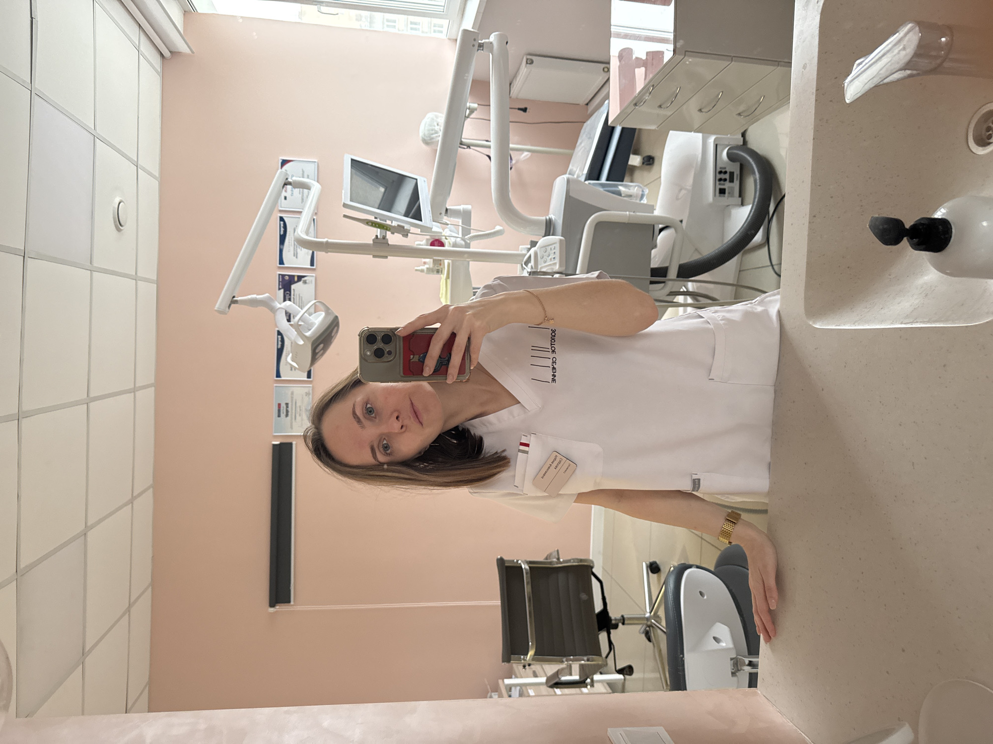 A woman is reflected in a mirror, holding a smartphone in her left hand to take a selfie. She is wearing a white medical uniform with a name tag and a logo that reads "ЗОЛОТОЕ СЕЧЕНИЕ" in Russian, suggesting a connection to a clinic of that name. The scene is inside a dental clinic, as indicated by the dental equipment in the background, including a dental chair, an overhead light, and a monitor. Certificates and documents are visible on the wall behind the dental equipment. Based on the logo on the uniform, the location is likely in the city of Perm, Russia, where a dental clinic called "Золотое Сечение" is located.