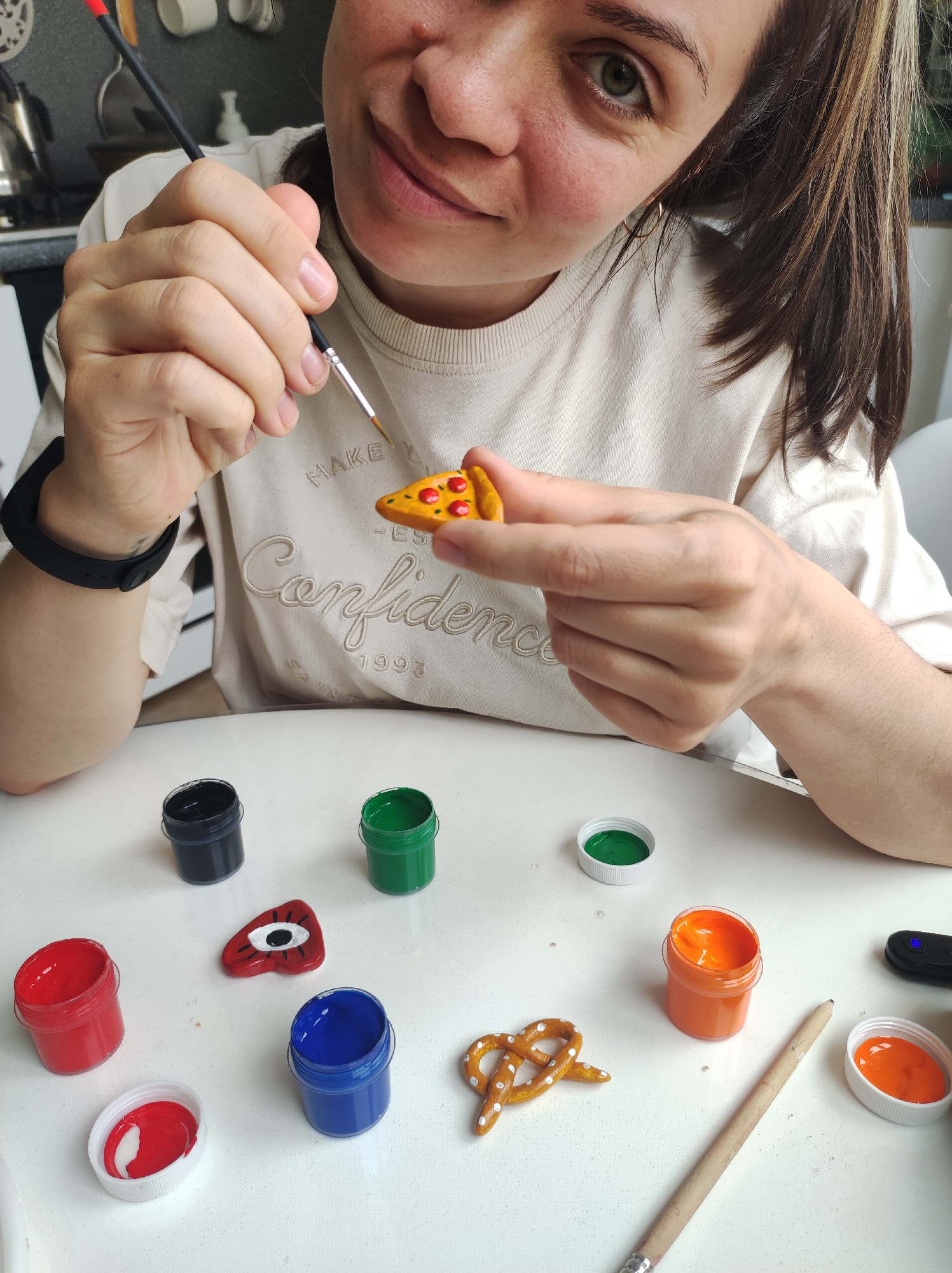 A woman with brown hair is painting a miniature pizza slice on a white table. She is using a thin paintbrush and the pizza slice is already painted yellow with red circles on top. Various paint pots are open on the table around her, including red, blue, green, black, and orange. Also on the table are a small charm painted as a blue and red eye, a pretzel charm, a paintbrush, and a black device. The woman is wearing a light-colored t-shirt with "Confidence 1993" stitched on the front. The location is possibly a home in the city of Chaikovsky, Russia, based on the prompt.