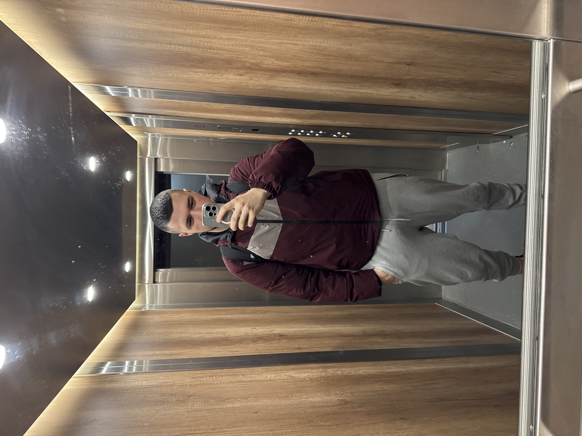 The image depicts a man in an elevator, reflected in a mirror. The man is holding a smartphone, presumably taking a picture of himself. He is wearing a maroon and grey jacket, and grey sweatpants. The elevator interior is a combination of wood paneling and metal, with a mirrored wall showing the reflection. There is a dark panel with lights in the ceiling. The location cannot be determined from the image alone, and there is no indication that it is in Sofia, Bulgaria.