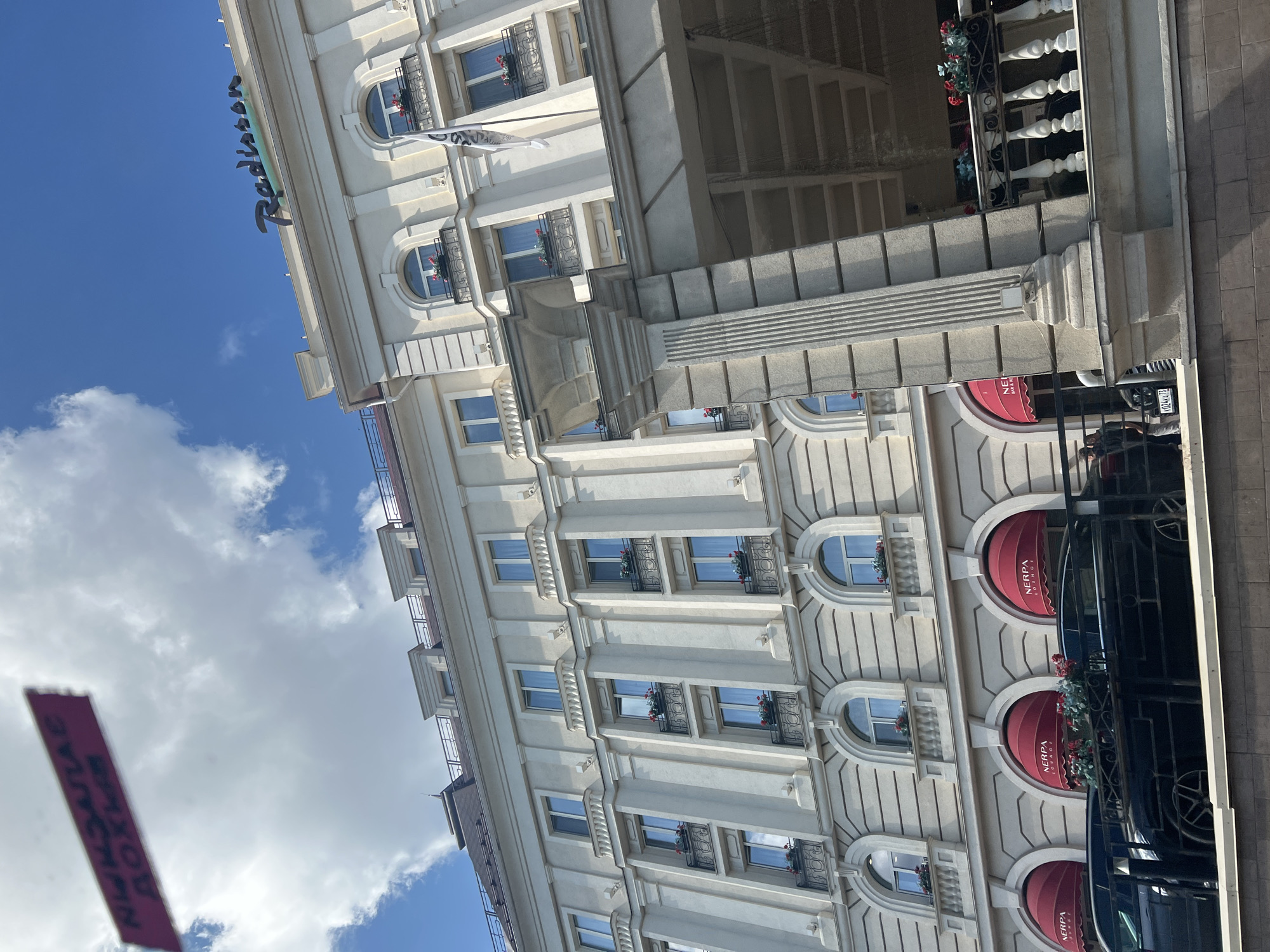 The image shows the Radisson Hotel in Ulyanovsk, Russia, on a sunny day with a blue sky and scattered clouds. The hotel building is light-colored, with multiple stories, arched windows, and balconies. The Radisson logo is visible at the top of the building. Red awnings with the text "NERPA LOUNGE" are present over some of the entrances. Two black cars are parked in front of the building. A red sign with blurry text is visible in the upper left corner of the image. The overall scene depicts the exterior of a hotel in an urban setting.