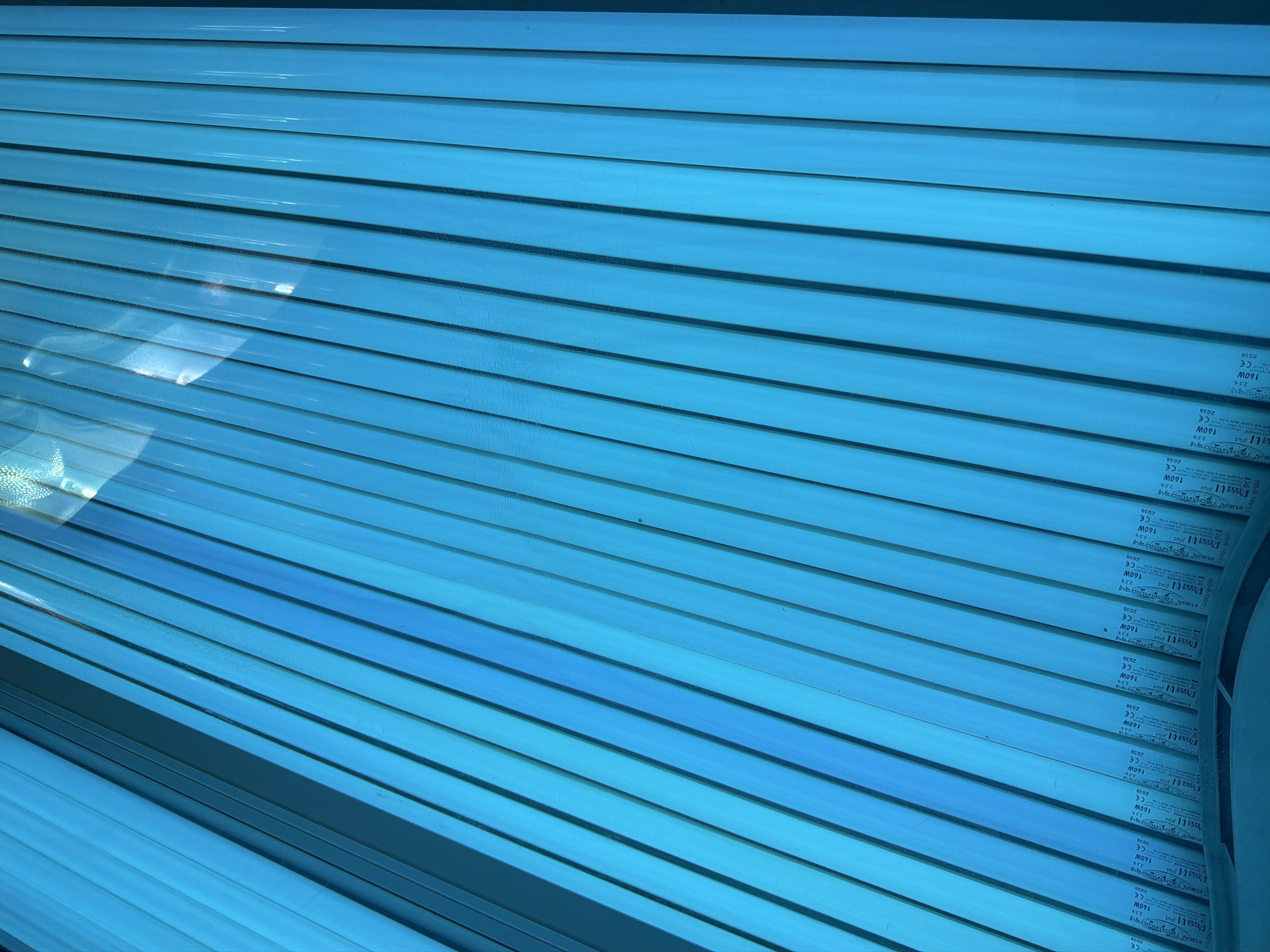 The image shows a close-up, diagonal view of a tanning bed, illuminated by blue fluorescent lights. The bed is composed of many parallel tubes, likely tanning lamps, arranged closely together. Some of the tubes have visible text printed on them, which includes "FASSA UI plus", "160W", "CE", "2039", and "3.5%". These details suggest that the tanning lamps are rated at 160 watts and have a specific UV percentage. The lighting creates a bright, somewhat sterile atmosphere. There are no people visible in the image. The scene is likely an indoor tanning salon, and the time of day and weather are not discernible from the image itself. The overall impression is one of a functional piece of equipment designed for cosmetic purposes.