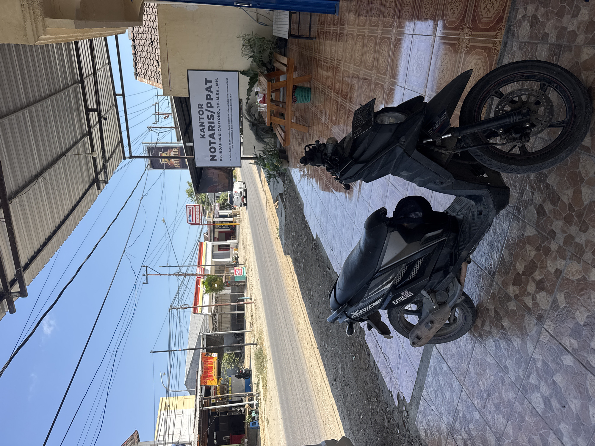The image captures a street scene in Taliwang, Indonesia, on a bright, sunny day. In the foreground, a black scooter is parked on a tiled patio. The scooter has "X-RIDE" and "EXTREME" branding visible on its body. To the right of the scooter, a wooden picnic table with benches sits under a covered patio. A green plastic basket is placed beneath the table. A white sign advertising "KANTOR NOTARIS/PPAT DR. INDAH DUGI CAHYONO, SH. M.Kn., MH." is mounted on the wall behind the table. The sign also displays contact information including an address on Jln. Sultan Syahrir, a phone number, and an email address.
Beyond the patio, a paved road stretches into the distance, lined with various buildings. A red and yellow "Alfamart" store is visible on the right side of the road, and further down, other shops and structures can be seen. The sky is clear blue with some scattered clouds. Overhead, power lines crisscross the sky. A white car is driving on the road in the distance. The overall environment suggests a commercial area in a town or city.