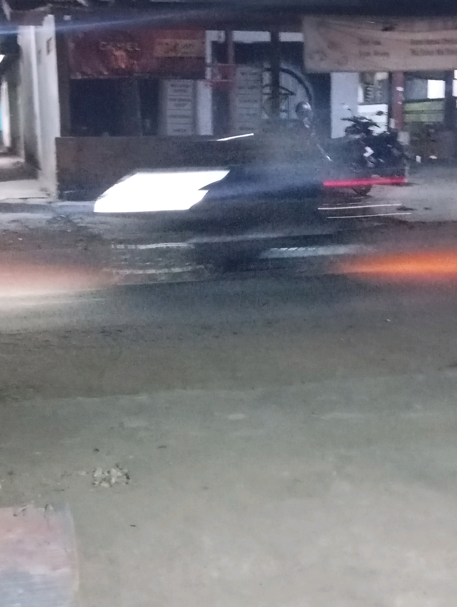 In Leman Benawi, Indonesia, the image captures a street scene at night, possibly in a commercial area. A motorcycle is prominent in the middle ground, its rear wheel emitting a bright red glow, suggesting it is either braking hard or has just accelerated, creating a reddish light trail on the wet asphalt. Smoke or exhaust fumes are visible around the motorcycle, obscuring some details. To the left of the motorcycle, a white reflective surface, possibly a piece of discarded material or a building feature, casts a bright glare on the ground. In the background, buildings with signs are partially visible, with one sign appearing to have text that includes "CAMEL". The overall lighting is dim, typical of nighttime conditions, with artificial lights from the buildings and the motorcycle contributing to the illumination. The wet road surface indicates recent rain or damp conditions.
