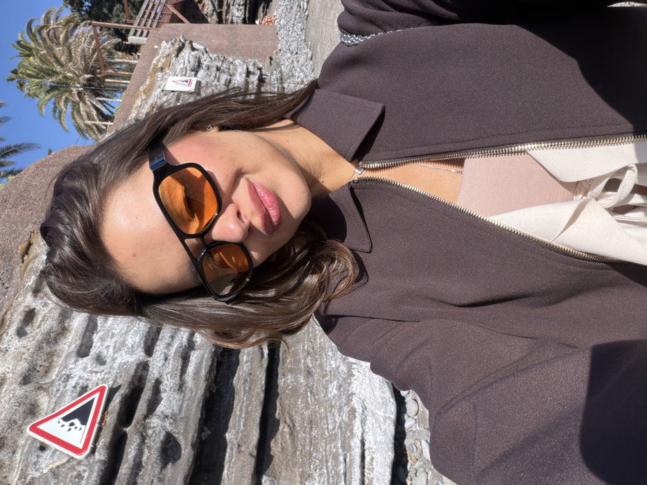 A selfie shows a person in a brown jacket with orange-tinted sunglasses lying on a gray, textured surface. Part of a white garment is visible under the open jacket. In the background, a palm tree and clear blue sky are visible. A triangular warning sign is situated in the bottom left of the frame, adding to the outdoor, possibly cliffside location. The person's face is angled towards the camera, emphasizing their lips and eyewear.