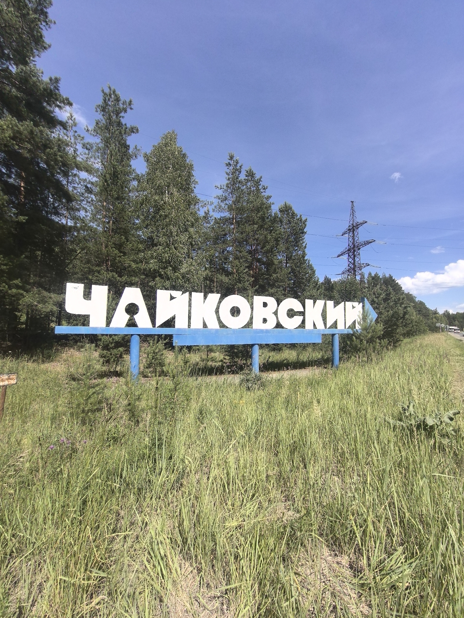 The image shows a white sign with blue supports and a blue arrow indicating direction. The sign has the Russian text "ЧАЙКОВСКИЙ" written in large white capital letters. It is situated in a grassy, open area with trees and vegetation in the background. A power line tower and electrical wires are visible in the distance, suggesting a rural or semi-rural setting. The sky is blue with some scattered clouds. The lighting indicates it is daytime, likely late morning or afternoon. The overall impression is a roadside sign in a natural landscape, possibly marking the entrance to a town or city.
