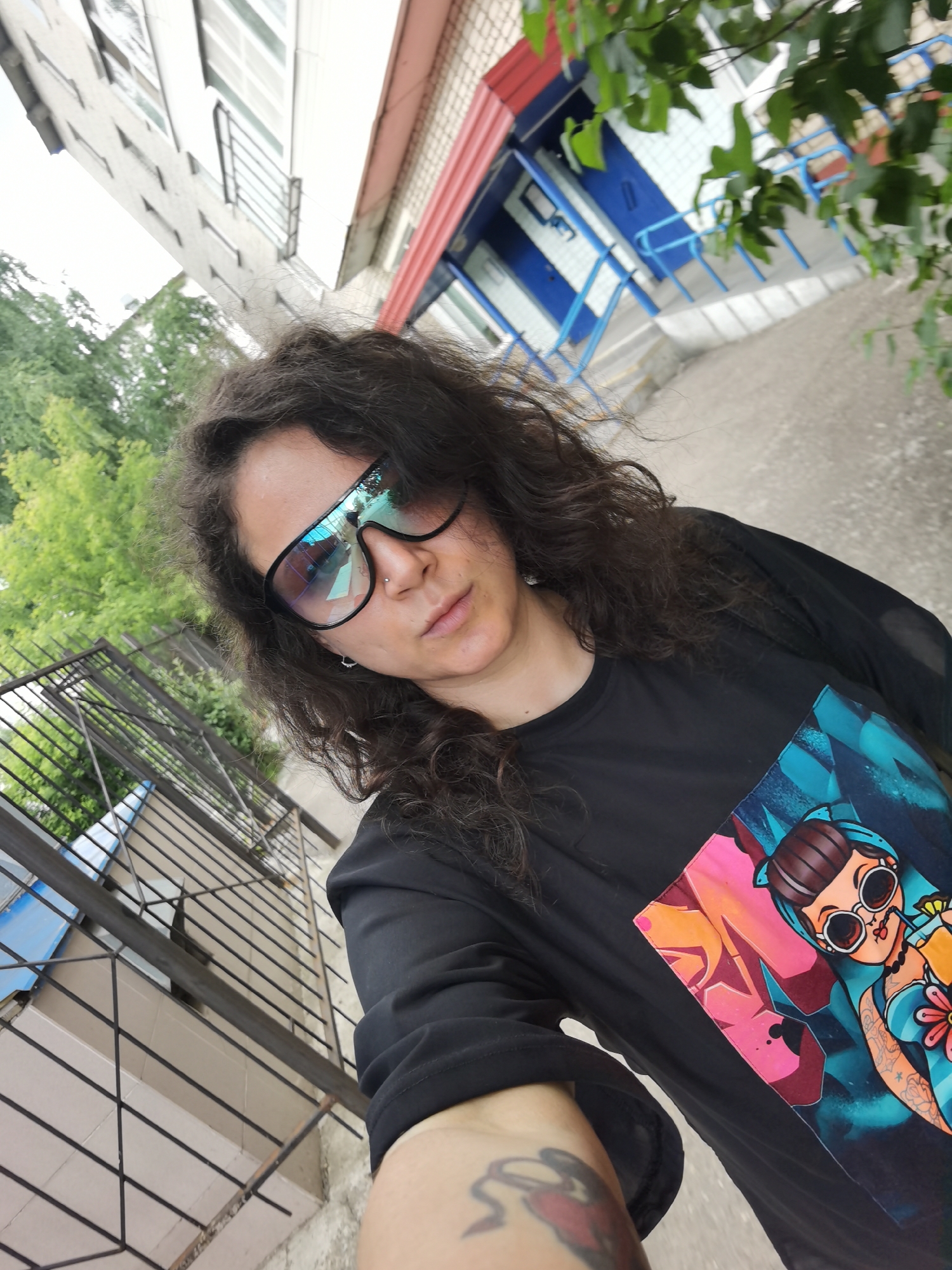 In a close-up, low-angle selfie, a person with dark, curly hair and sunglasses is positioned in the foreground. They are wearing a black t-shirt with a colorful graphic of a stylized female cartoon character. The person's right arm, bearing a tattoo, is visible.
The background suggests an outdoor urban setting in Chaikovsky, Russia. To the left, a tall, light-colored building with numerous windows and dark window bars is visible, angled upwards. To the right and behind the subject, a building entrance with a red-topped overhang, blue railings, and blue and white striped walls is partially obscured by green foliage. A metal fence or railing runs diagonally across the lower left portion of the image. The ground appears to be paved. The overall lighting indicates it is daytime, with a slightly overcast sky.