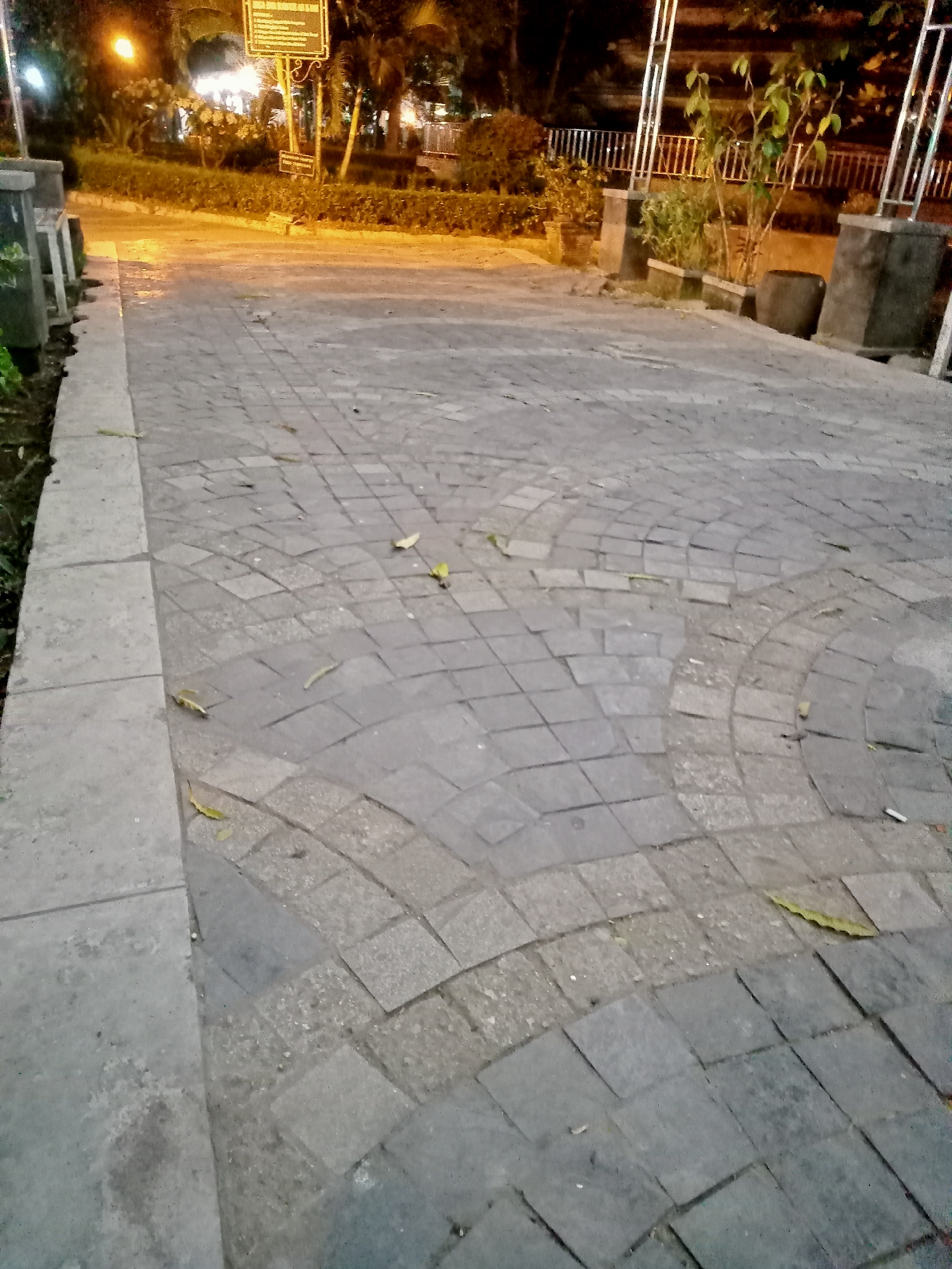 The image depicts a pathway paved with square gray tiles, some of which are arranged in a curved pattern. The pathway is wet, suggesting recent rain or cleaning. Scattered leaves are visible on the tiles. On the left side of the pathway, there is a concrete border and some greenery. In the background, there is a dimly lit park with trees, bushes, and streetlights. A sign with text is mounted on a pole. The scene appears to be taken at dusk or at night. This pathway is likely located in a public area within the city of Tulungagung, Indonesia, possibly a park or a pedestrian walkway.