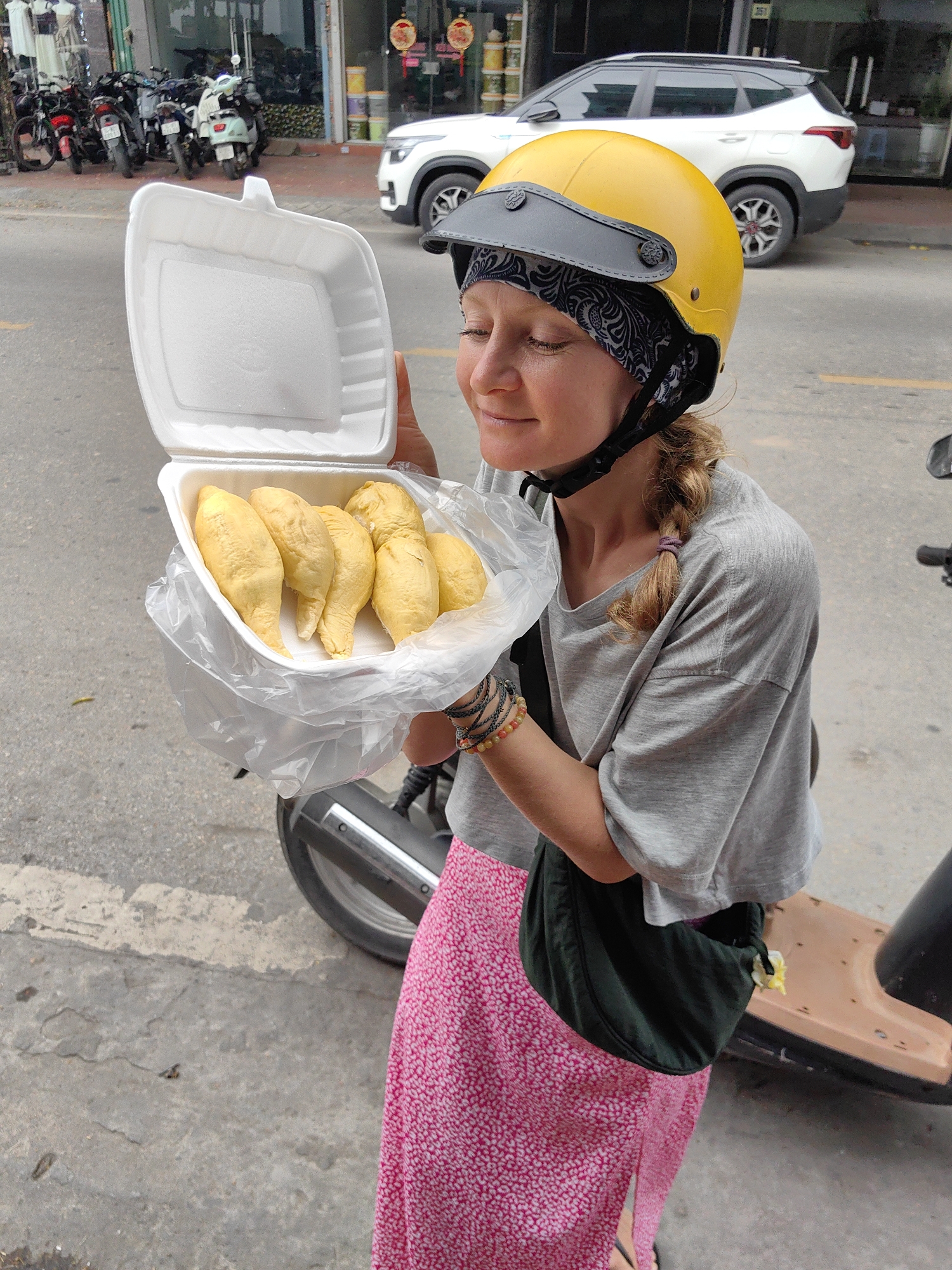 A woman wearing a yellow open-face helmet and a patterned bandana is captured outdoors, holding an open white styrofoam container. Inside the container, nestled within a clear plastic bag, are several vibrant yellow segments of peeled durian fruit. Her eyes are closed, and a gentle smile plays on her lips, suggesting she is savoring the aroma or anticipating the taste of the fruit. She is dressed in a grey short-sleeved t-shirt, a long pink skirt with a white pattern, and a dark sling bag across her body. Stacked bracelets are visible on her right wrist.
The scene takes place on a paved street in Cao Xanh Ward, Vietnam, during the daytime, likely under an overcast sky. In the background, several motorbikes and scooters are parked along the curb. A white SUV is also visible further down the street. Behind the parked vehicles, there are storefronts of a building, one with glass displaying items like clothing. Faint red circular decorations and an indistinct building number (possibly "251" or "257") are visible on the building's facade. A brown and black scooter is partially visible in the lower right, next to the woman. The image conveys a moment of pleasure and anticipation in an everyday Vietnamese street setting.
