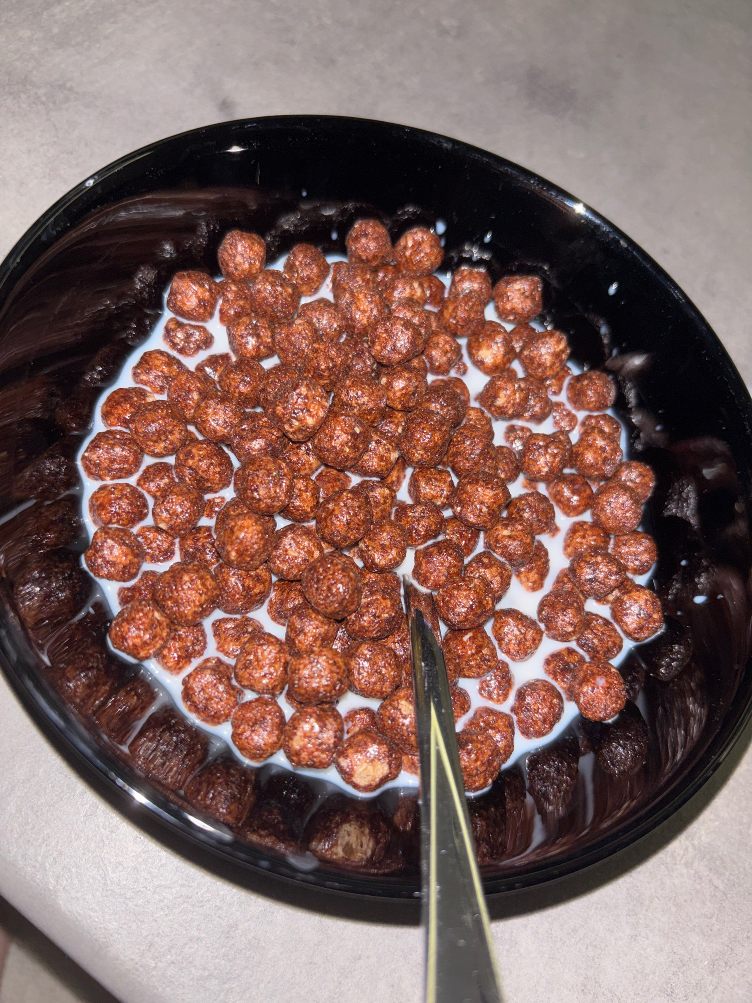 A black bowl filled with chocolate puffed cereal and milk sits on a textured, grey surface. A silver spoon is partially submerged in the cereal, its angled head visible. The cereal pieces are clustered together, with milk seeping between them and around the edges of the bowl. The lighting in the image is dim, creating reflections on the glossy surface of the bowl. There is no discernible location in the city of Ruse, Bulgaria.