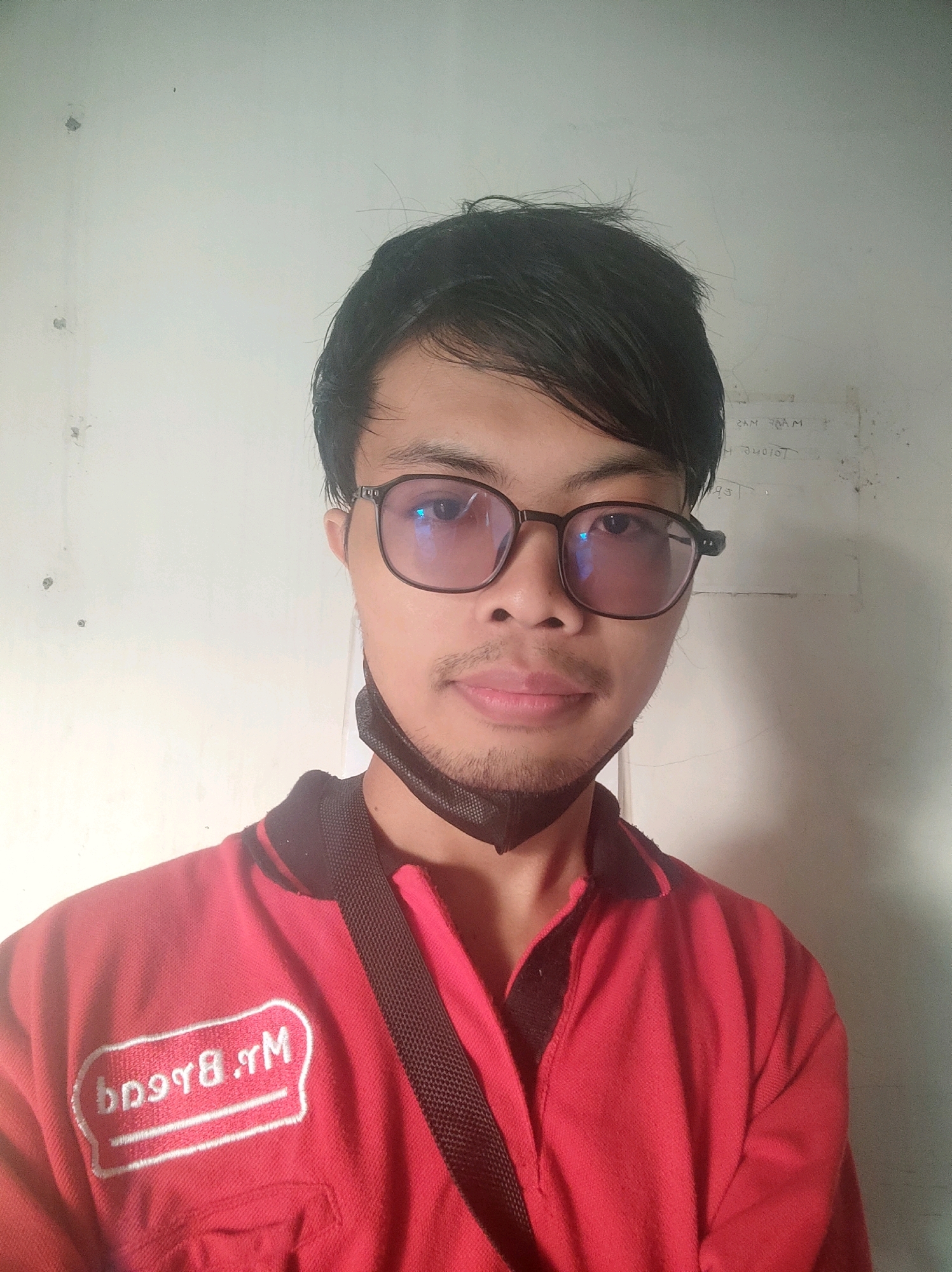 A selfie of a young East Asian male wearing glasses and a red shirt with a black collar is shown. The man has black hair and is wearing a black face mask around his neck. A white oval patch with red text is visible on the left side of the shirt. The background is a plain, off-white wall. The image was taken indoors.