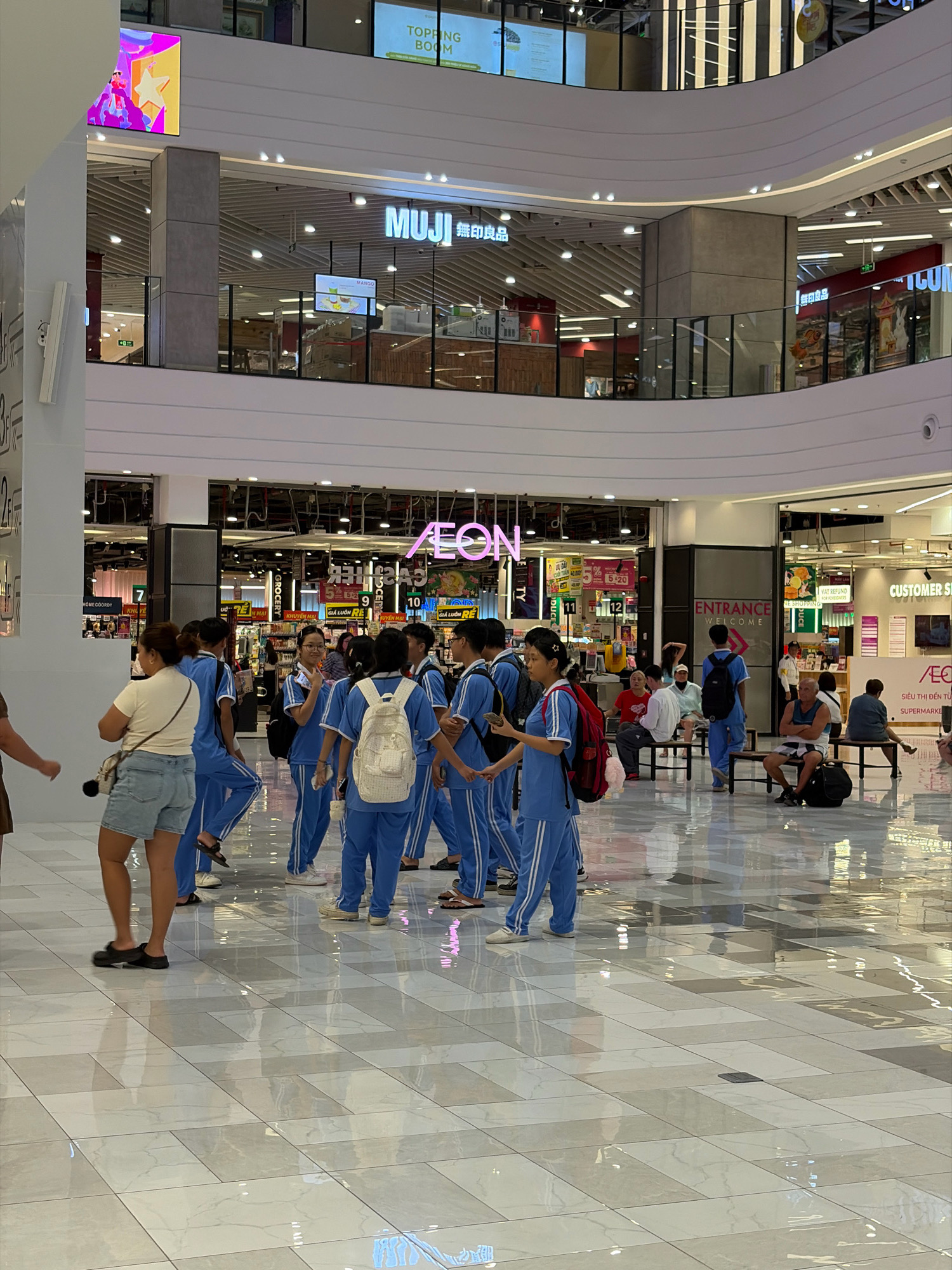 The image shows the interior of a modern shopping mall in Da Nang, Vietnam, likely at the AEON Mall. In the foreground, a group of young people in matching blue uniforms, possibly students on a field trip or sports team, are gathered in the open concourse. Some are looking at their phones, while others are interacting with each other. One person in the foreground is a woman wearing a light-colored t-shirt and denim shorts, carrying a crossbody bag. Behind the group, various shops and storefronts are visible, including "MUJI" and "AEON SUPERMARKET". The mall features a multi-level design with escalators and walkways. The floor is made of polished, light-colored tiles that reflect the overhead lighting. Several other individuals are seated on benches in the background, observing the scene or waiting. The overall atmosphere appears to be one of casual activity and shopping.