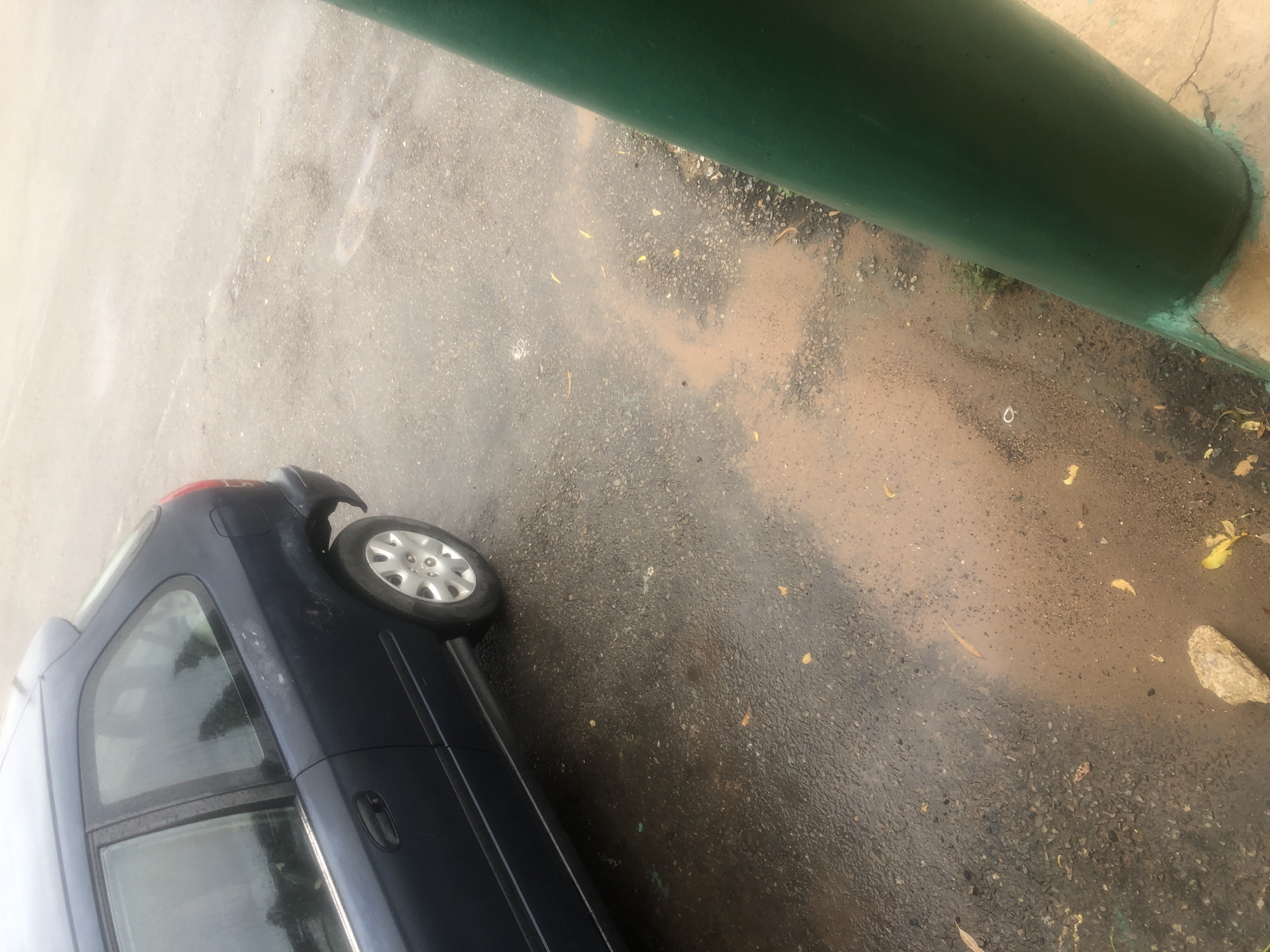 An eye-level, medium shot captures the rear driver's side of a dark blue, five-door hatchback parked on a wet asphalt road, possibly in Jalingo, Nigeria, given the context. The car is angled slightly away from the viewer. The vehicle's rear bumper is missing, exposing some of the underlying structure. The road surface is dark and wet, with visible puddles and streaks of reddish-brown mud, indicating recent rain or a spill. Fallen yellow and brown leaves are scattered across the asphalt. To the right of the frame, a large, dark green cylindrical pole is positioned diagonally, with its base resting on a cracked concrete surface. The pole is partially obscuring the road and a patch of sparse green grass growing at its base. The overall lighting is overcast, suggesting a cloudy or rainy day. There are no people or animals visible in the scene.