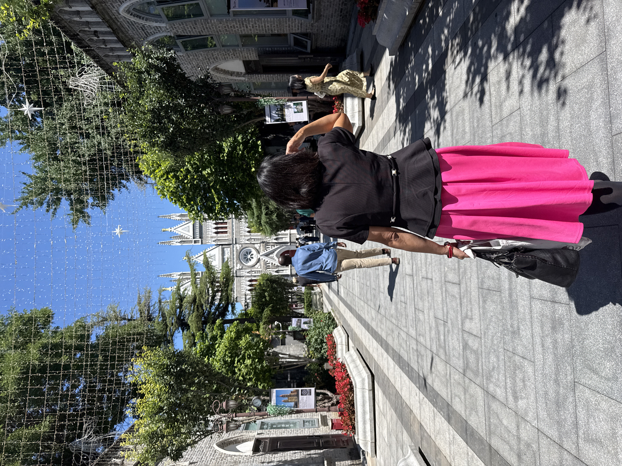 The image depicts a paved pedestrian walkway in Xicheng District, Beijing, China, leading towards the Xishiku Church (North Church) in the background. The church is a large, Gothic Revival structure with multiple spires, a prominent rose window, and light-colored stone facade with red accents. The walkway is lined on both sides by two-story buildings constructed of grey brick with arched windows and doors.
In the foreground, a woman is seen from behind, walking away from the camera. She wears a black short-sleeved peplum top with a thin white-accented belt and a bright pink midi skirt. She carries a black handbag with a white bag inside in her left hand, which also features a red bracelet. Her right hand is raised towards her head. Ahead of her, a man walks in the same direction, wearing a light blue long-sleeved shirt and tan trousers. To the right, another woman walks diagonally towards the foreground, wearing a yellow-patterned dress and carrying a light-colored handbag. Several indistinct figures are visible further down the path.
The walkway is paved with grey rectangular tiles, featuring a central strip of darker grey tiles. Large green trees line the path, casting distinct shadows on the ground, indicating bright sunlight. Smaller green bushes and rectangular planters containing red flowers are arranged along the sides. Overhead, a grid-like structure of wires spans the walkway, from which numerous small, reflective or light-emitting strands hang. Several white star-shaped ornaments and a larger white wire-frame angelic figure are suspended from this overhead network. Ornate black lampposts are visible, with one on the left displaying a clock and information boards. The sky is clear and blue.