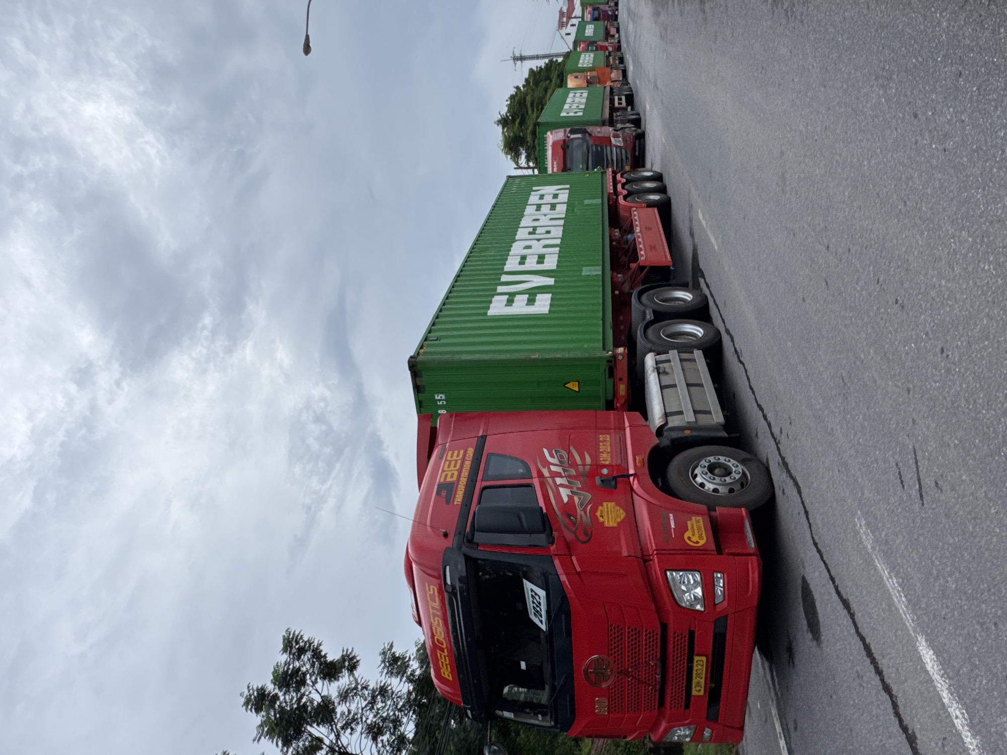 In a scene captured in Lao Bảo Commune, Vietnam, a convoy of trucks, predominantly red with green shipping containers, lines a paved road. The lead truck, a bright red vehicle, displays "BEELOGISTICS" on its upper fascia and the "BEE TRANSPORTATION CORP" logo on the side of its cabin. The green shipping container on this truck is emblazoned with the prominent "EVERGREEN" logo. Following closely behind, other trucks, also carrying "EVERGREEN" containers, extend into the distance, suggesting a logistics operation underway. The sky above is overcast with a mix of light and dark clouds. Trees and vegetation are visible on the left side of the road, and in the distance, a power pole and some buildings can be discerned. The road surface shows some wet patches and tire marks. The overall impression is one of organized transport activity in a semi-urban or rural setting.
