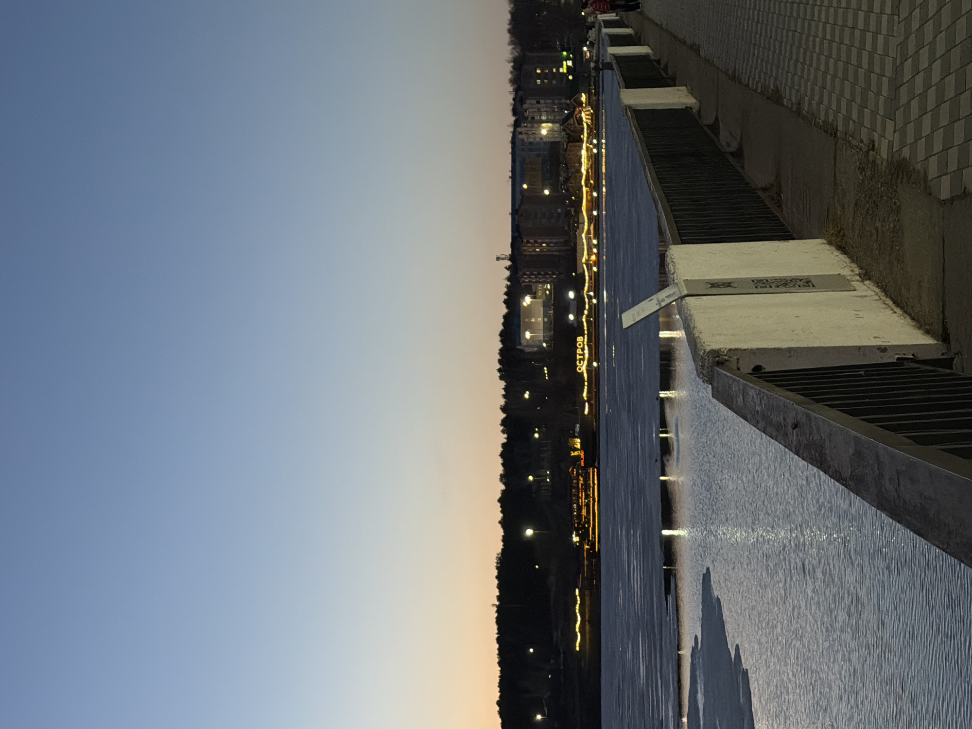 The image depicts an evening view of the Izhevsk Pond embankment in Votkinsk, Russia. The sky transitions from blue to orange, suggesting twilight. The embankment's stone and metal barrier runs along the right side of the image, with the pond filling the central space. Lights from the cityscape reflect on the water's surface. Across the pond, distant buildings and trees are faintly illuminated by streetlights. A sign "ОСТРОВ" (OSTROV - Island) is partially visible on the opposite shore. The tiled pavement is on the top right corner.