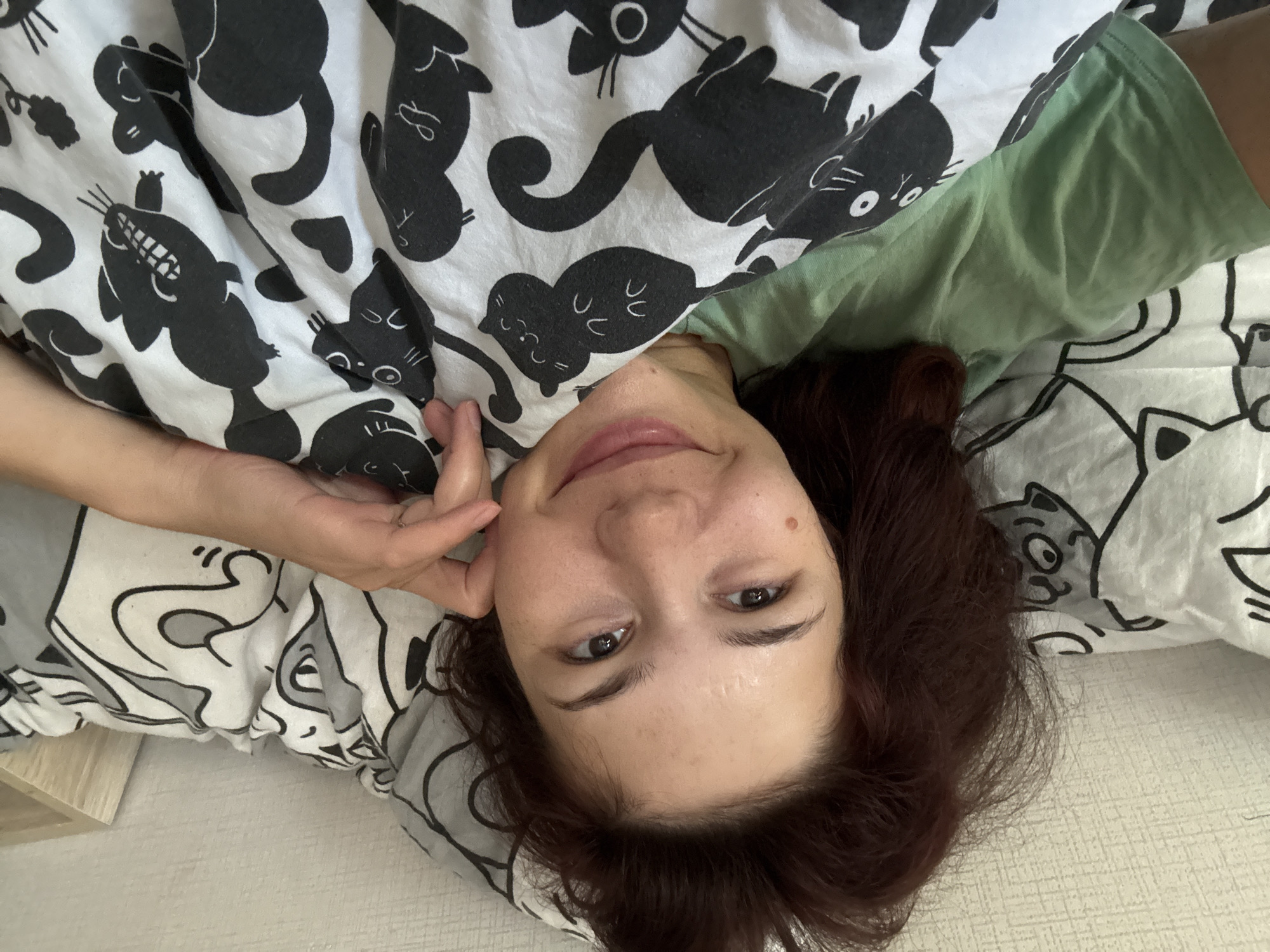 A young woman with brown hair is lying in bed, looking at the camera. She is wearing a green t-shirt and is covered by a blanket with a black and white cat print. The room appears to be a bedroom, with a wall and a headboard visible in the background. The lighting suggests it is daytime.