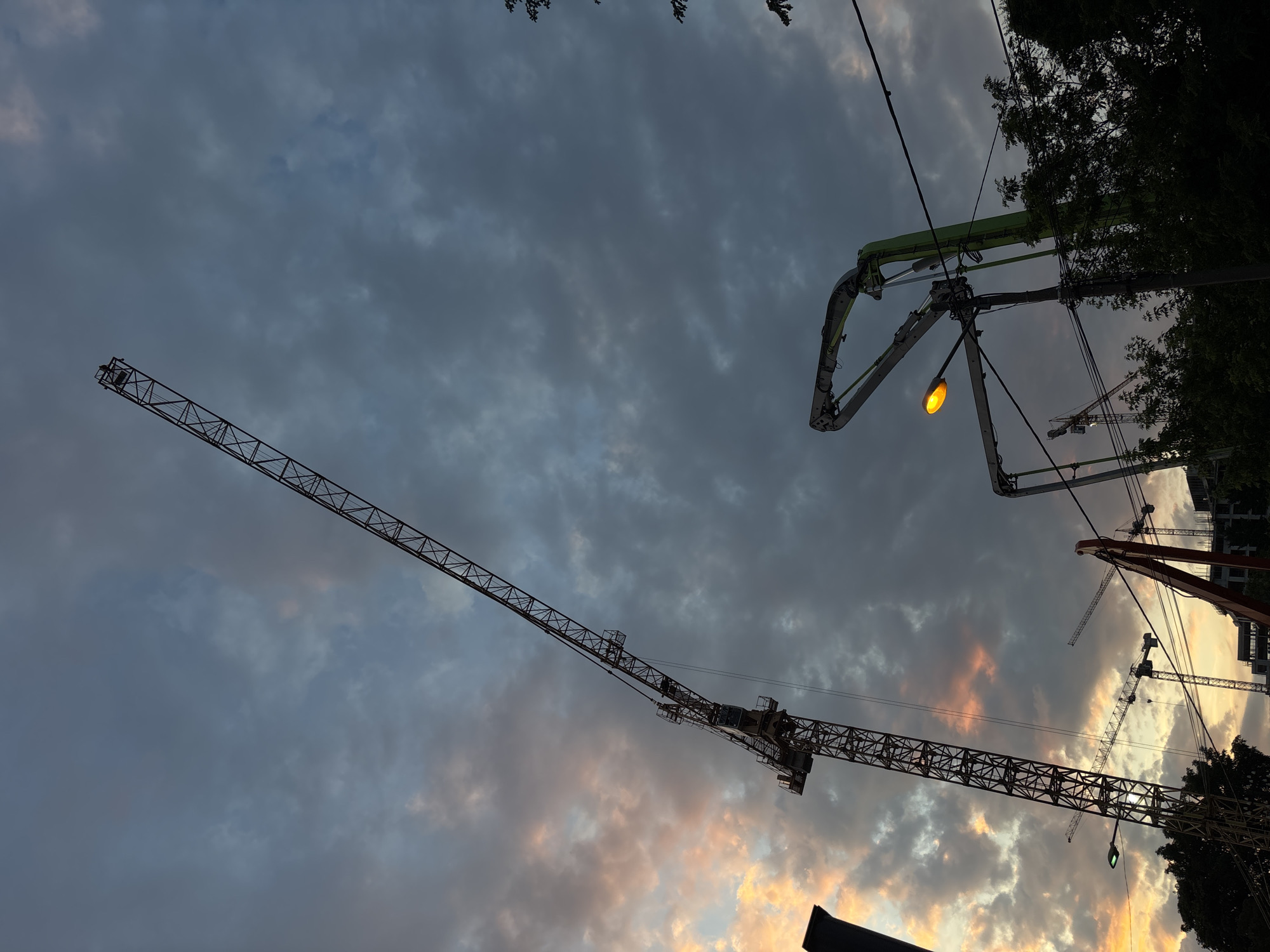 The image depicts a dusk or dawn scene in Belgrade, Serbia, characterized by a cloudy sky with hues of gray, blue, orange, and pink. The foreground shows multiple construction cranes, indicating an active construction site. The silhouette of trees is visible on the right side of the image. Overhead power lines intersect the view, adding to the urban landscape. A concrete pump with an extended boom is visible at the top.