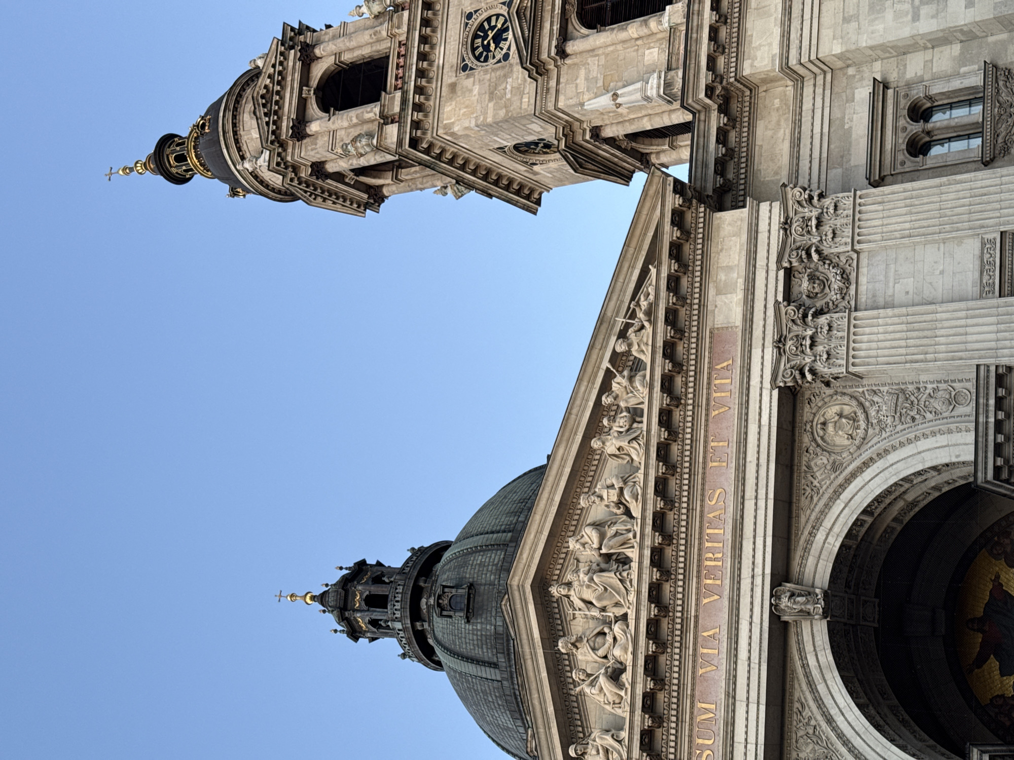 This image shows the architecture of St. Stephen's Basilica in Budapest, Hungary. The camera is pointed upward, capturing the dome and one of the towers. The dome features intricate carvings and a smaller tower on top. The tower, also ornately decorated, has a clock on its face. The sky is clear and blue in the background. Inscriptions are visible on the building's facade.