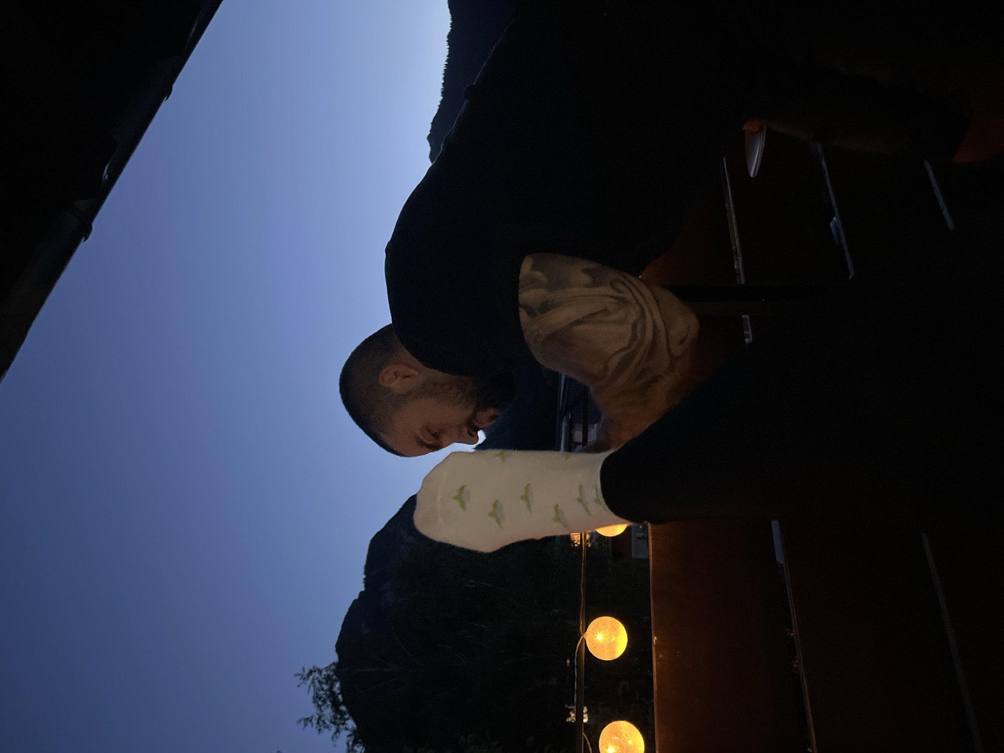 At dusk, two people are on a balcony overlooking the Bavarian Alps in Garmisch-Partenkirchen, Germany. A man with a beard and tattoos on his arm is leaning forward, facing a woman's leg that is raised and facing the camera. She is wearing a white sock with green whale patterns and black leggings. In the background, there are mountains, trees, and string lights that emit a warm orange glow. The sky is a deep, clear blue, transitioning into night. The balcony railing is made of metal, and a dark overhang is visible at the top of the frame.