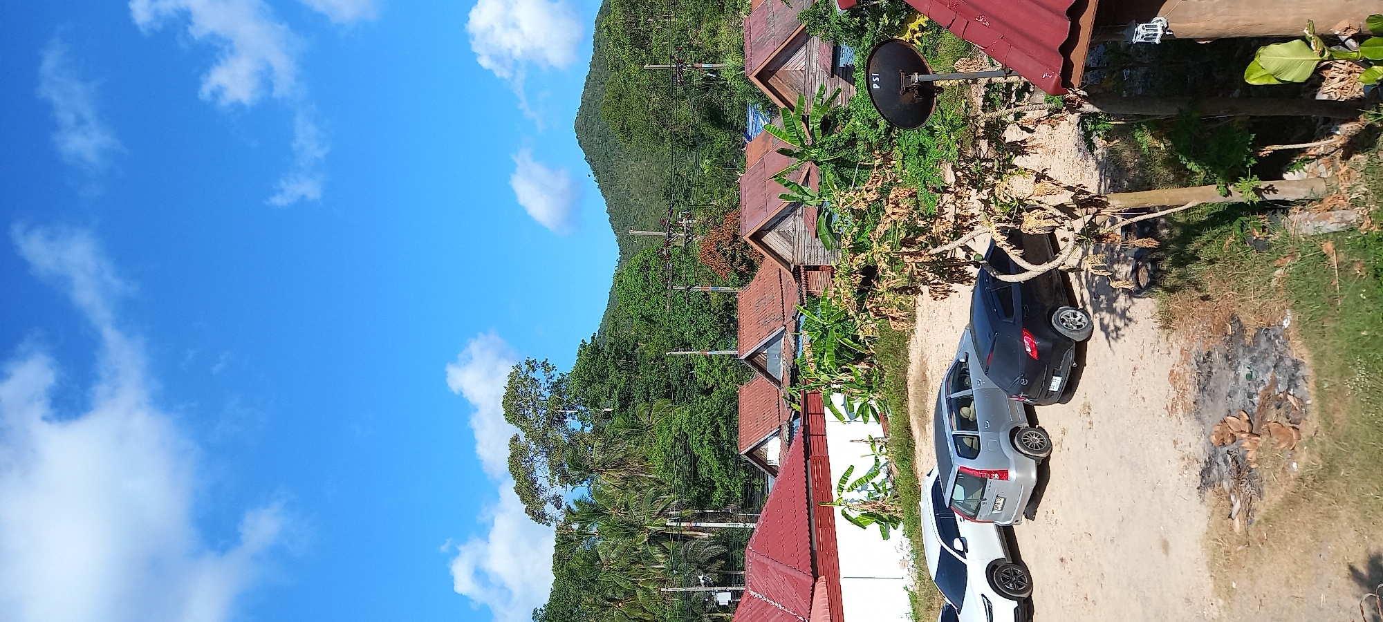 This photograph captures a vibrant daytime scene in Baan Chaloklum, Thailand, depicting a village or residential area nestled against a lush, green mountain range. The sky is a clear, brilliant blue dotted with scattered, fluffy white clouds, indicating fine weather.
In the mid-ground, several small, rustic bungalows or houses are visible, featuring pitched roofs made of reddish-brown corrugated metal. The structures appear to be made of wood, blending into the natural surroundings. Abundant tropical vegetation, including tall trees, palm trees, and banana plants, surrounds the dwellings and extends up the steep mountainside in the background.
The foreground consists of an unpaved, dirt road or ground area where three cars are parked. A silver SUV or crossover is prominently featured, with a dark grey or black sedan parked behind it. The front portion of a white car is partially visible at the bottom of the frame. Near the parked cars and houses, a black satellite dish with the text "PSI" emblazoned on it is mounted on a pole. Several utility poles with overhead power lines traverse the scene, indicative of local infrastructure. Patches of dry grass and some scattered debris, possibly burnt material, are visible on the ground to the right of the cars.
No people are visible, suggesting a quiet moment in the village. The bright sunlight suggests it is mid-day or afternoon, contributing to the serene and peaceful atmosphere of the tropical setting.