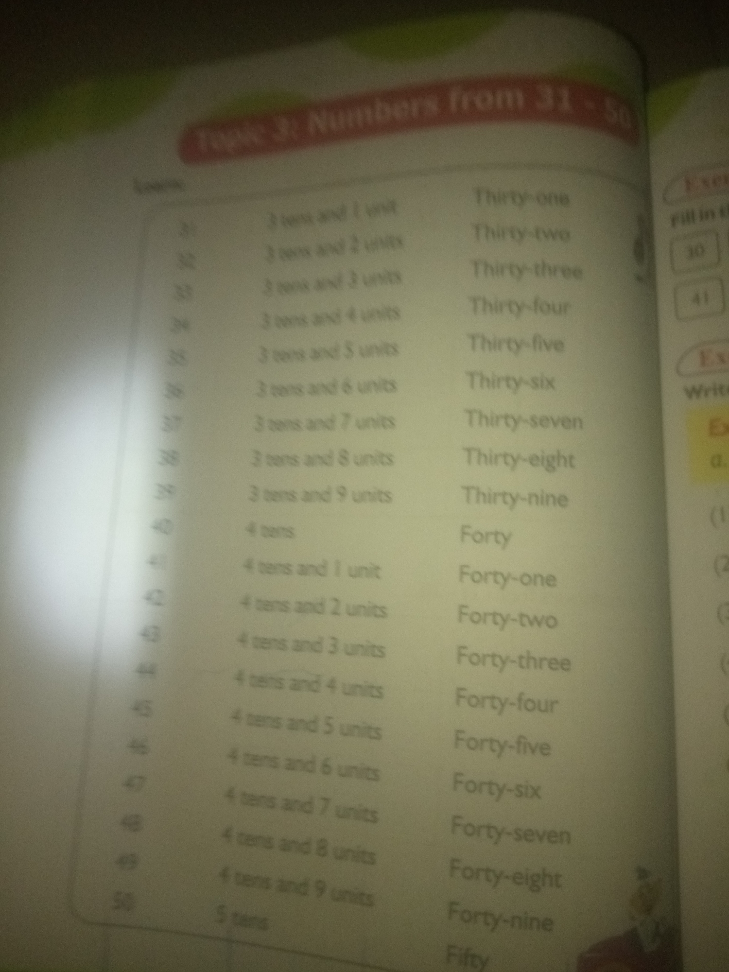 The image is a close-up of a page from a book, likely a children's educational book, displaying a list of numbers and their corresponding word forms. The page has a title "Topic 3: Numbers from 31 - 50". On the left side, there is a column of numbers from 31 to 50. To the right of these numbers, there are two columns of text describing the numbers: one column with a format like "3 tens and 1 unit" and the other column with the written-out form of the number, such as "Thirty-one". There is also a section on the right side of the page that appears to be an exercise or activity, with the heading "Exer" and some instructions like "Fill in the" and "Write". The lighting in the image is uneven, with a bright spotlight effect originating from the left, obscuring some details on that side of the page. The background is blurry and appears to be the interior of a room. There is no information in the image that relates to the city of Jauro Yinu, Nigeria.