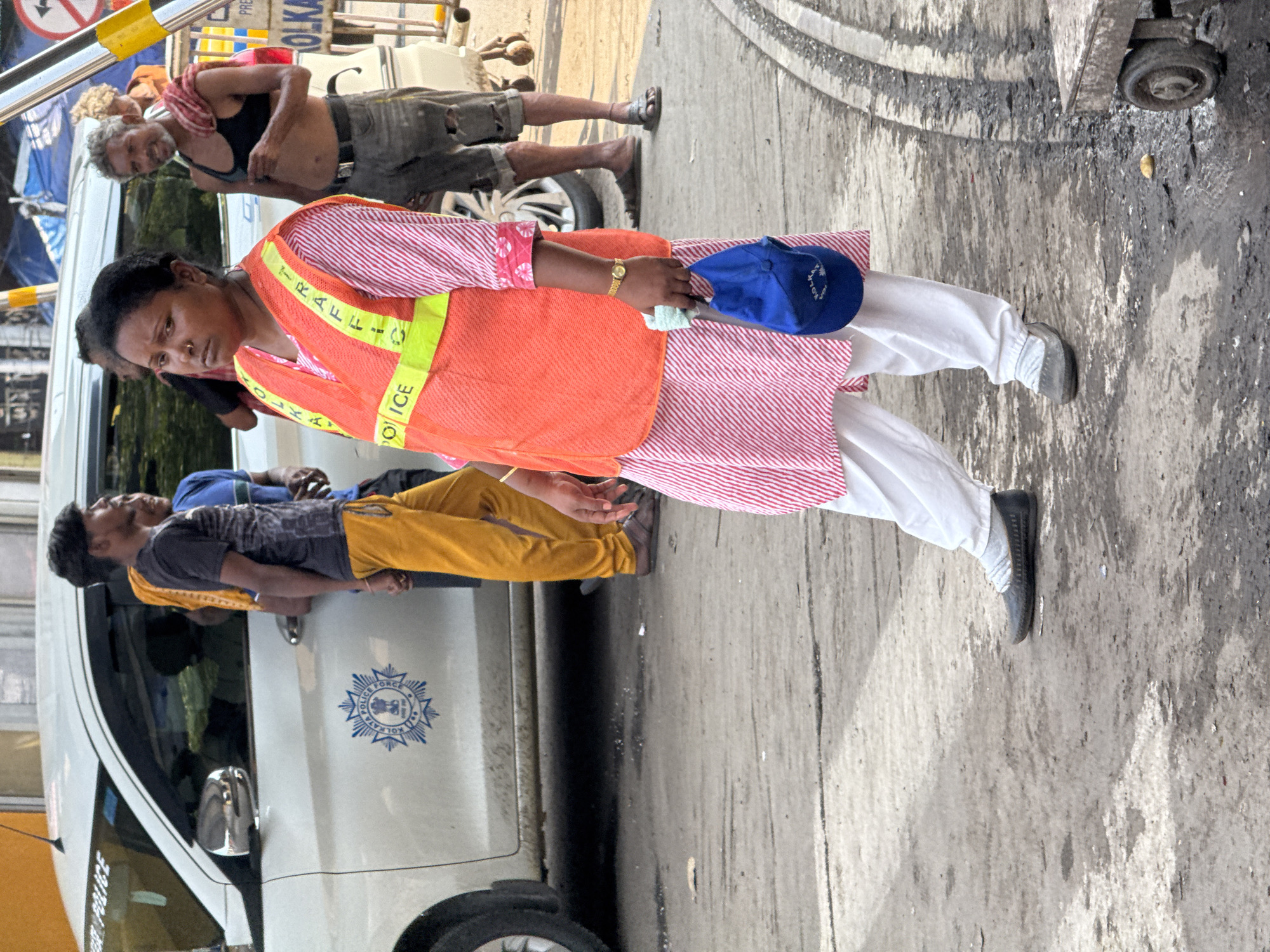 The image shows a street scene in Kolkata, India. A South Asian woman wearing a red and white striped dress, an orange traffic vest, white pants, and black shoes stands on a gray concrete surface, holding a blue baseball cap. Behind her are three men, two leaning against a white vehicle marked with "KOLKATA POLICE FORCE" crest, and a shirtless man standing further back. The environment includes parts of vehicles, possibly for crowd control, and a dirty concrete ground with cracks and stains.