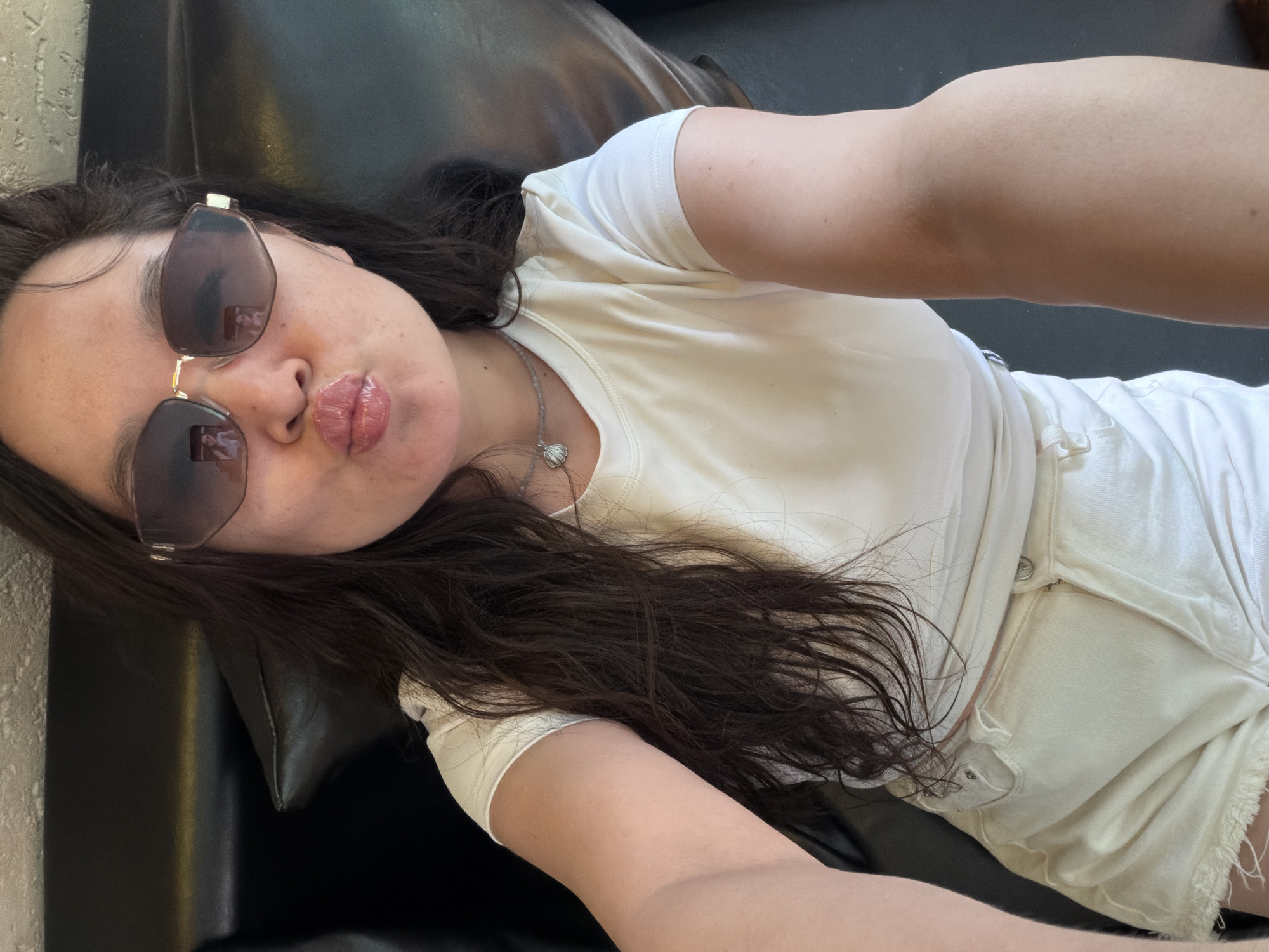A woman lies on a black leather sofa. She is wearing sunglasses, a white t-shirt, and white shorts. She has long dark hair and is wearing a necklace with a pendant. The location appears to be indoors.