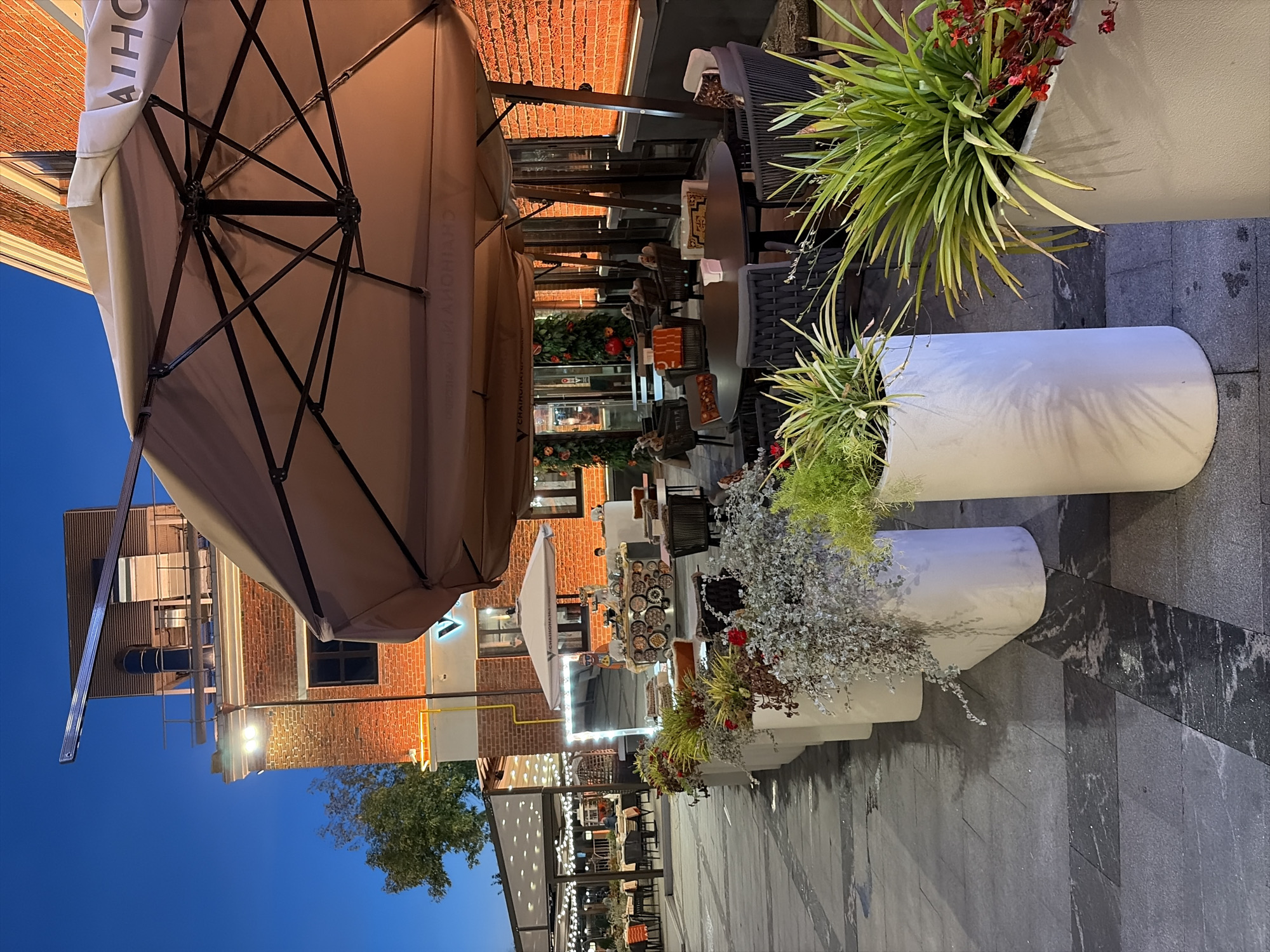 The image depicts an outdoor seating area of a restaurant or cafe located in Moscow, Russia, during twilight. A large, beige umbrella is prominently featured, casting a shadow over a circular table surrounded by dark wicker chairs with patterned cushions. In the foreground, several large white planters contain lush green plants and some red flowers. Behind the seating area, a brick building with illuminated windows and architectural details is visible. Further in the background, another covered seating area with string lights is set up, with tables and chairs. The overall ambiance suggests a pleasant evening setting for dining or socializing.
