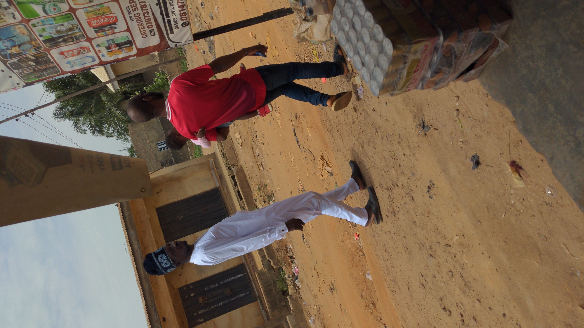 The image depicts an outdoor scene in Ado, Nigeria. In the foreground is a man dressed in a white traditional outfit with a patterned cap. Further back, a man in a red shirt and jeans is holding a child. They are standing on a dirt road. A catering services advertisement board is visible, displaying various drink brands like Coca-Cola, and phone numbers. Adjacent to the advertisement, packages of canned goods stacked in a box. Buildings are visible in the background.