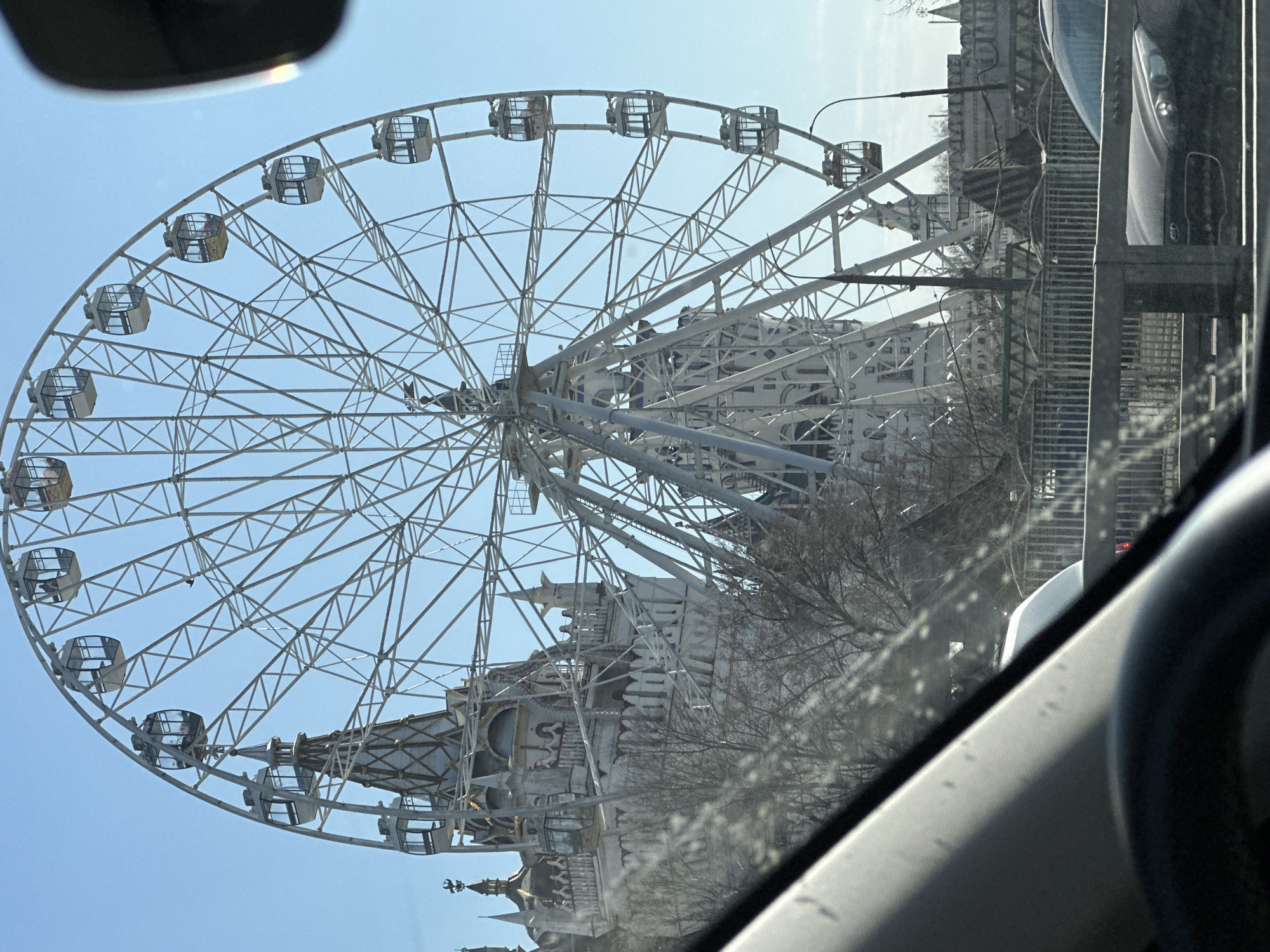 The image shows a large Ferris wheel in Moscow, Russia. The Ferris wheel is white and has enclosed gondolas. In the background, there is a building with ornate architectural details. The photo was taken from inside a car, as parts of the vehicle are visible in the foreground. The sky is clear and blue. Bare trees are visible near the Ferris wheel and building.