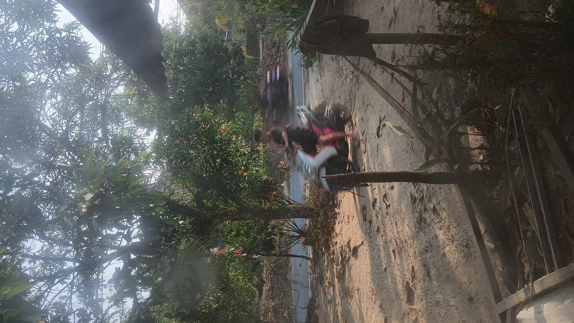 In the city of Prabumulih, Indonesia, a young man is seen riding a scooter on a dirt path. He is wearing a dark t-shirt and shorts. The scooter is white and red. In the background, there is a road with another scooter in motion. The scene is set amidst lush greenery with trees providing shade. The ground is covered with sand and fallen leaves, indicating an outdoor environment, possibly a rural or semi-urban area. The light suggests it is either early morning or late afternoon.
