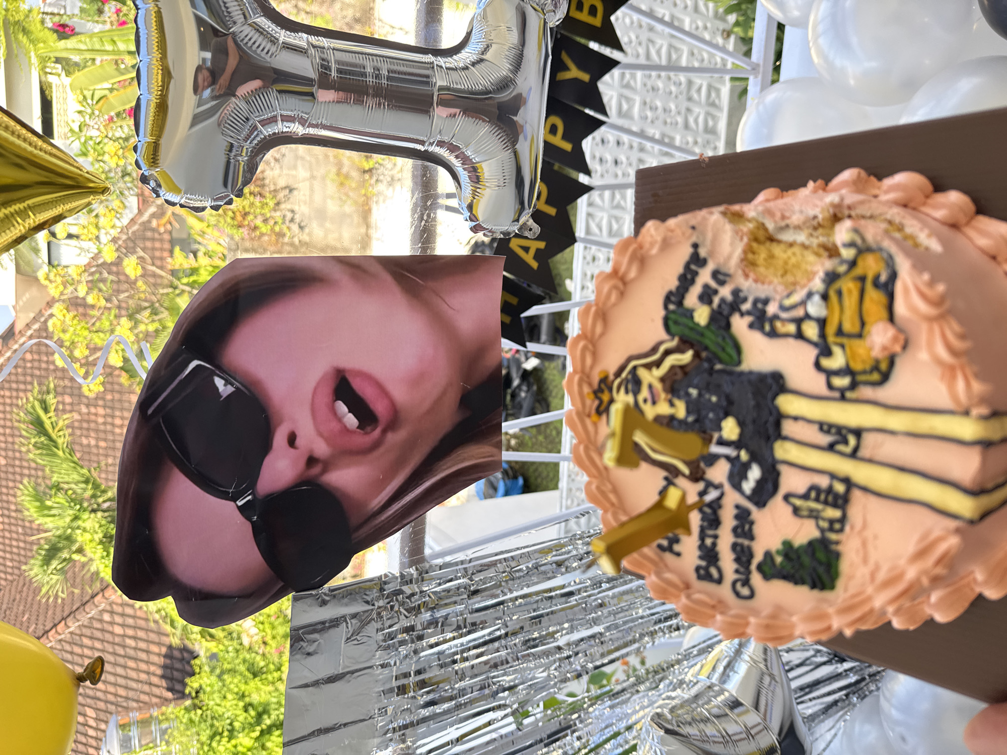 The image depicts a birthday celebration with a cake and decorations. The cake is round and frosted in a peach color, decorated with a drawing of a cartoon character holding a shopping bag and standing next to a yellow car. The cake has the words "Happy Birthday Queen" written on it in black frosting. There are two gold number candles, one shaped like a "7" and another like a "1." To the left of the cake, there are silver fringe curtains. In the background, a large foil balloon shaped like the number "71" is visible. A cutout of a person's face with sunglasses and an open mouth is also present. A black banner with yellow letters spells out "HAPPY B" and part of "Y B" hangs behind the cake. Several white balloons are scattered in the foreground. The scene appears to be outdoors, with greenery and building rooftops visible in the background, suggesting a location in North Kuta, Indonesia.