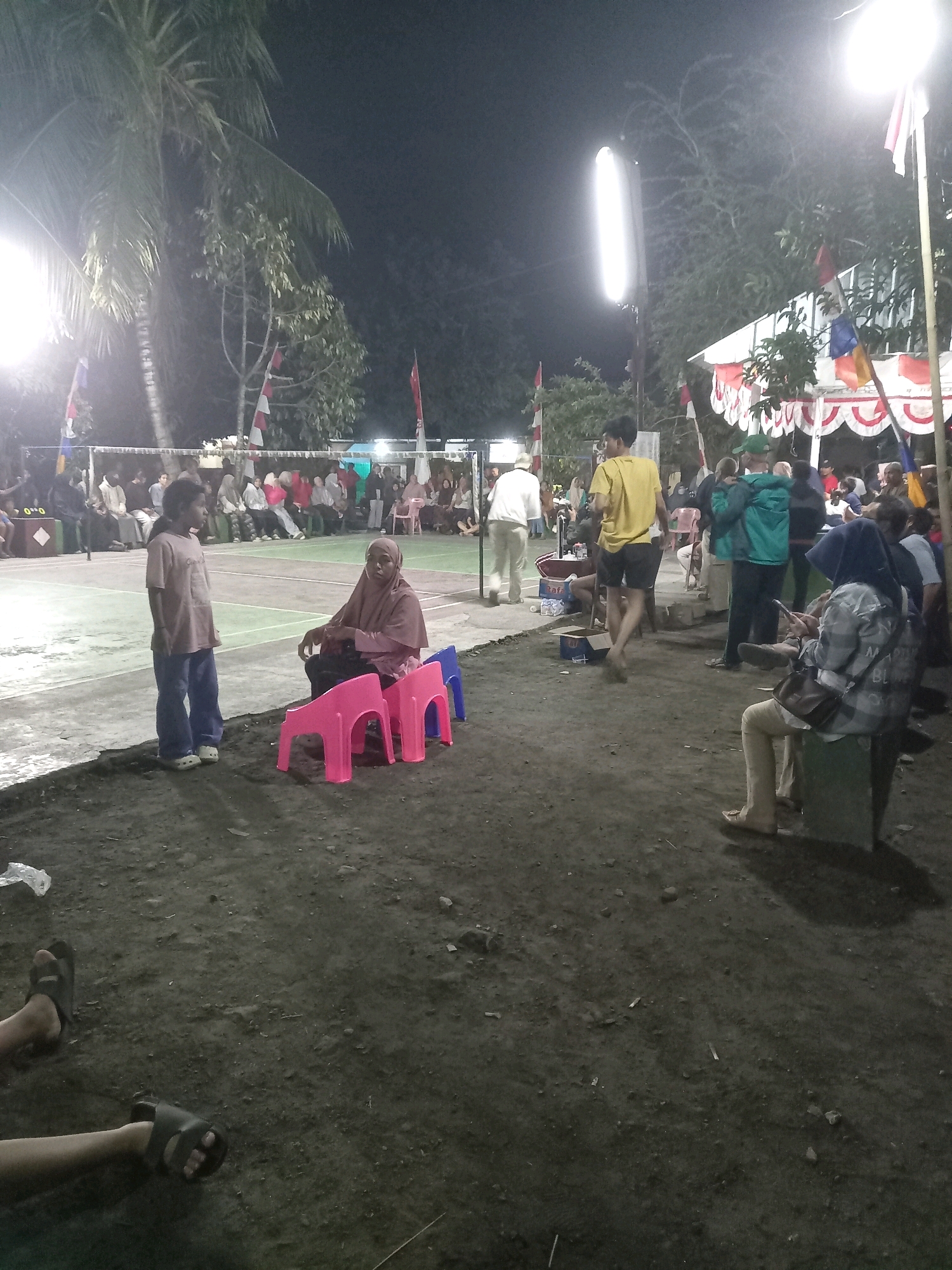 The image depicts an outdoor gathering at night, likely in Banyumulek, Indonesia, with a volleyball court illuminated by bright lights. In the foreground, a young girl stands on the edge of the court, looking to her right, while a woman in a headscarf sits on a pink plastic chair nearby, also looking in the same direction. Behind them, a crowd of people are seated on benches, watching an event. The scene is further enlivened by Indonesian flags and decorative banners. The ground is a dark, rough terrain, with scattered debris. The overall atmosphere suggests a community event or sporting activity taking place after dark.