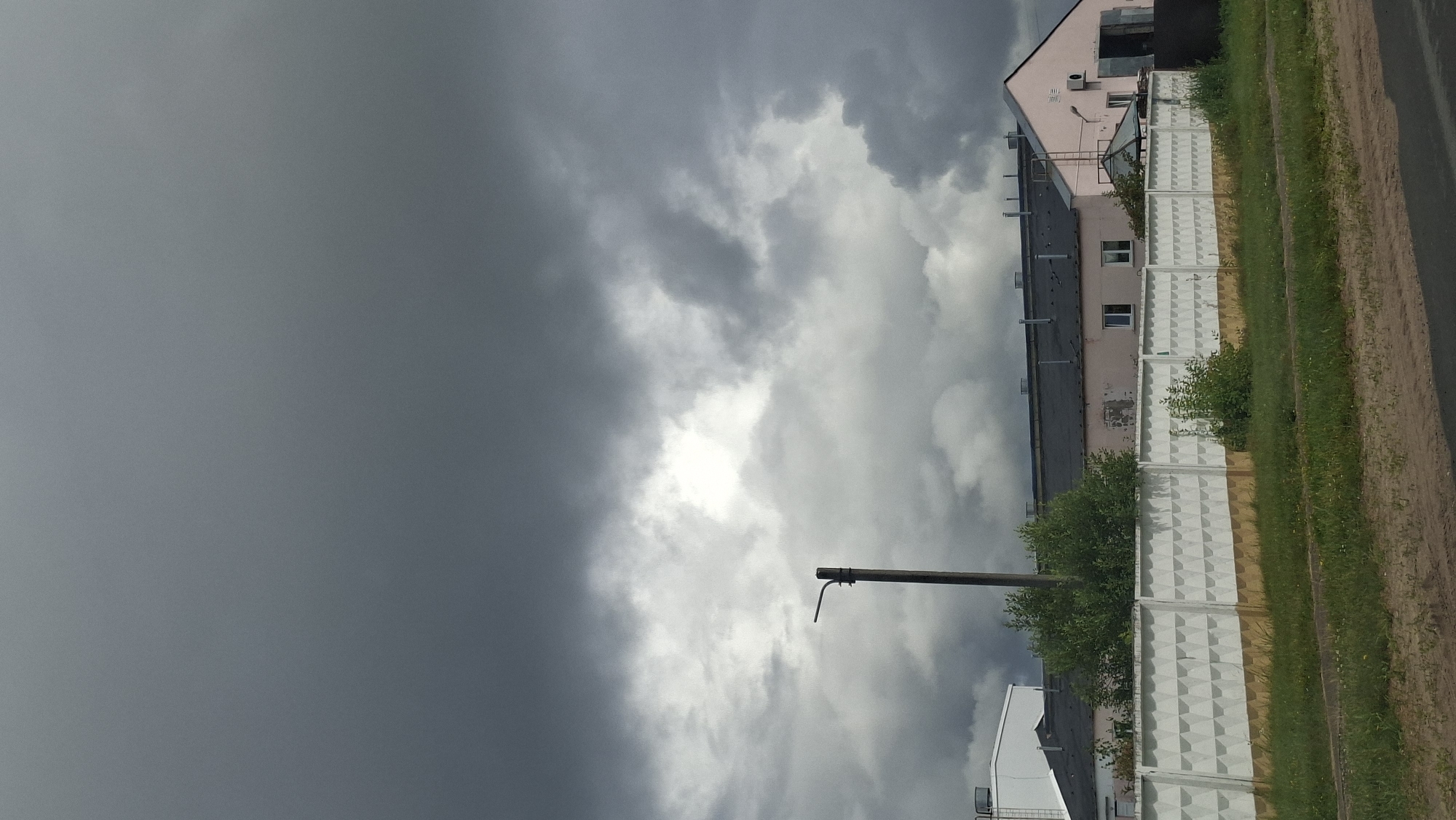 The image depicts an overcast sky with dramatic, dark clouds gathering over a light-colored, flat-roofed industrial building. A light post stands prominently in the middle ground, silhouetted against the stormy sky. In the foreground, a white concrete fence with a patterned surface runs horizontally across the scene, separating the industrial area from a strip of green grass and a dirt road. The industrial building is long and appears to be a warehouse or factory, with several windows visible on its side. The overall impression is one of an impending storm in an industrial or rural setting. The specific location in Myadzyel, Belarus cannot be determined from the visual information provided.