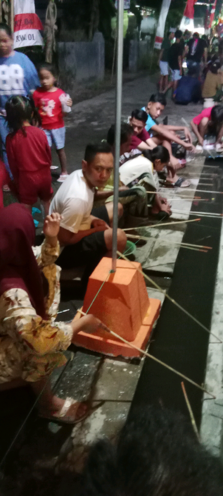 In Kesamben, Indonesia, at night, several people are gathered outdoors near a road. In the foreground, a woman in a maroon hijab and patterned dress is seated on the ground, facing a bright orange, tiered object. She appears to be interacting with this object, which is surrounded by thin wooden sticks. To her left, another person is partially visible.
Further back, a man in a white t-shirt and black shorts is sitting and looking towards the viewer with a neutral expression. He is positioned in front of a group of other individuals. Some are sitting on the ground, while others are standing. The standing individuals appear to be mostly young men and boys. One young boy in a blue shirt is visible in the middle ground, leaning forward.
Behind the seated individuals, a young girl in a red shirt and blue shorts is holding a small white cup. To her left, a woman in a blue t-shirt with "88" printed on it is walking away from the camera. There are also red and white banners or flags visible in the background, suggesting a possible event or celebration. The ground appears to be paved and somewhat wet or dirty. The lighting is dim, indicating it is likely evening or night. The overall scene suggests a community gathering or activity taking place outdoors.